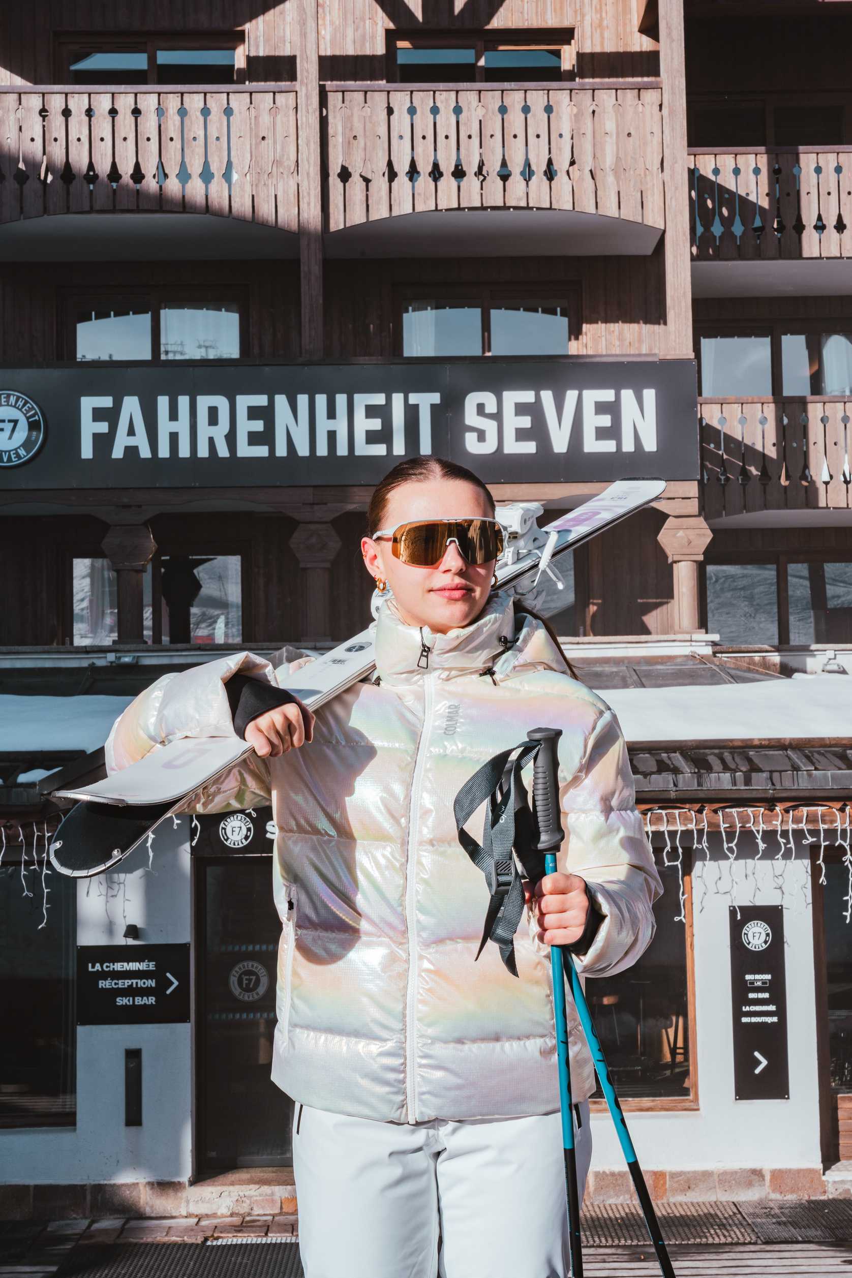 Jeune femme en tenue de ski irisée et lunettes de soleil tenant des bâtons de ski et portant des skis sur son épaule, devant un bâtiment en bois avec une enseigne indiquant Fahrenheit Seven.