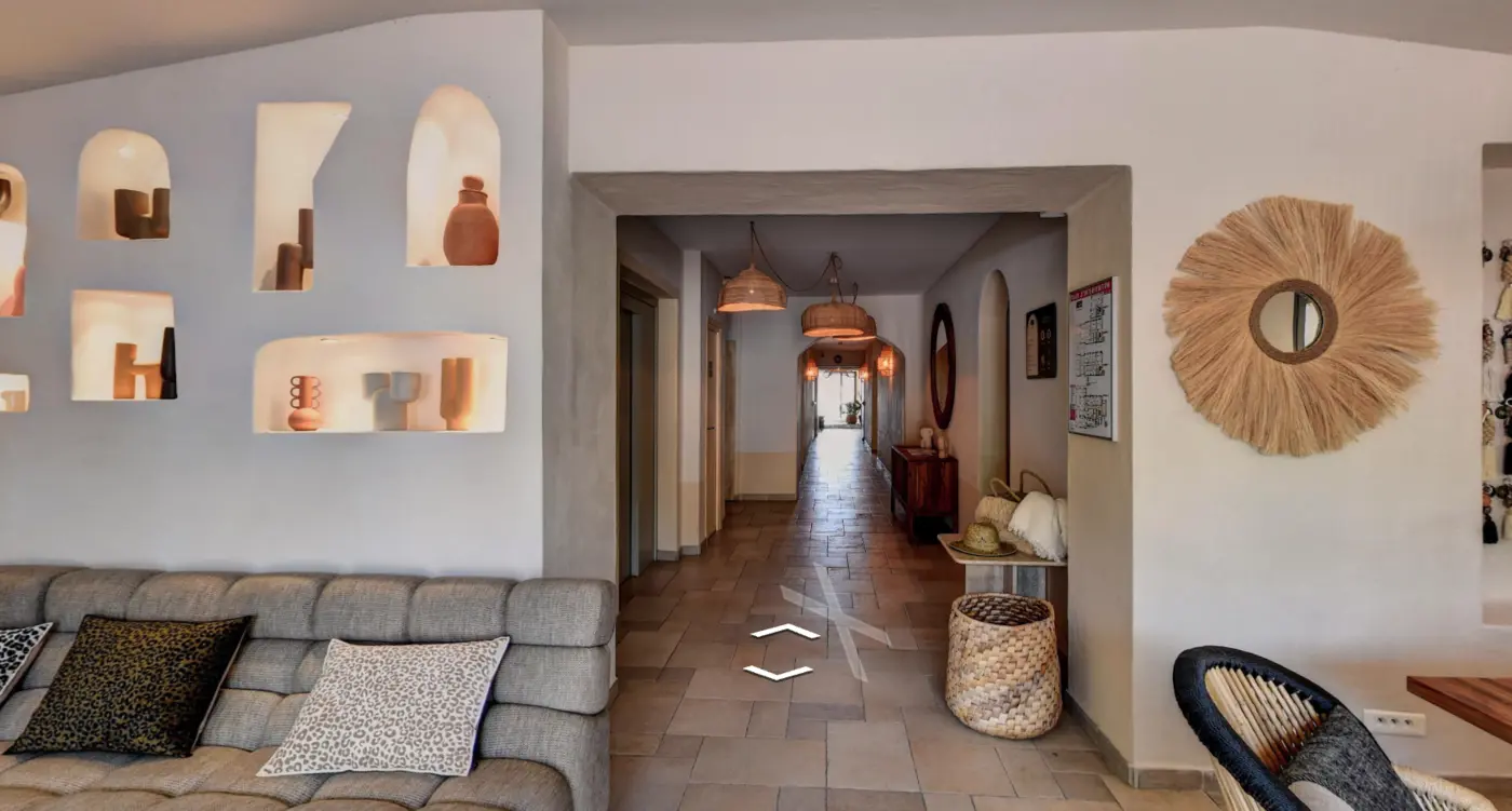 Intérieur d'un couloir d'hôtel ou appartement avec mur blanc, canapé gris avec coussins à motifs léopard, niches murales éclairées décorées de pots et objets, miroir rond encadré de fibres naturelles, mobilier en bois et suspensions en osier, ambiance chaleureuse et moderne.