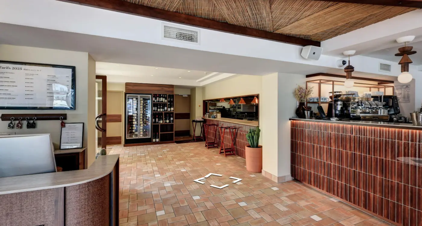 Intérieur lumineux d'un café ou bar avec comptoir en bois carrelé à droite, machines à café, tabourets en bois contre un bar au fond avec lampes suspendues et cave à vin encastrée au mur, sol en carreaux de terre cuite, plafond avec poutres et matériaux naturels.