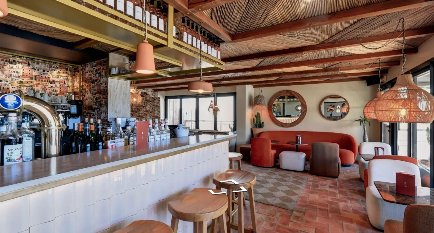 Intérieur élégant d'un bar avec un comptoir blanc sur la gauche, des tabourets en bois, diverses bouteilles alignées, un plafond avec poutres en bois et tissage naturel, des sièges colorés orange et beige autour de petites tables, deux grands miroirs ronds décoratifs au mur, et des suspensions lumineuses en osier.