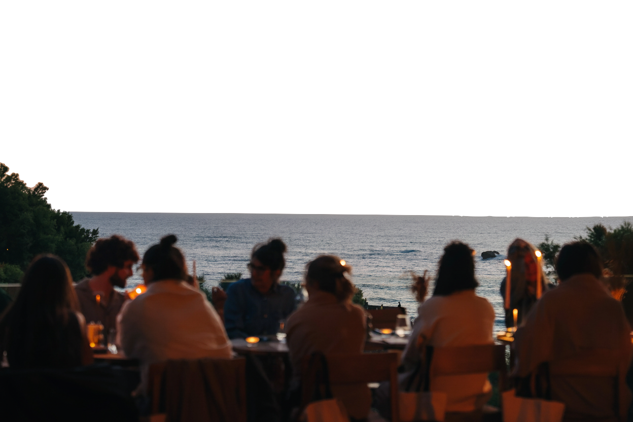 Un groupe de personnes assises autour d'une table en plein air au crépuscule, avec des bougies allumées, face à une mer calme et un paysage de végétation à l'arrière-plan.