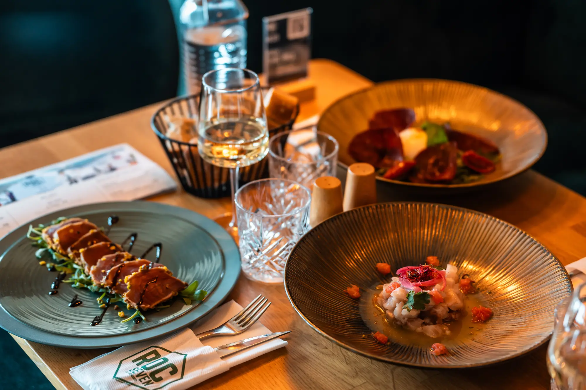 Table with three gourmet dishes: sliced seared tuna on greens drizzled with sauce, a seafood tartare garnished with herbs and pickled onions, and a plate with slices of cured meat and a lit candle, accompanied by two crystal glasses, a glass of white wine, and a bread basket.