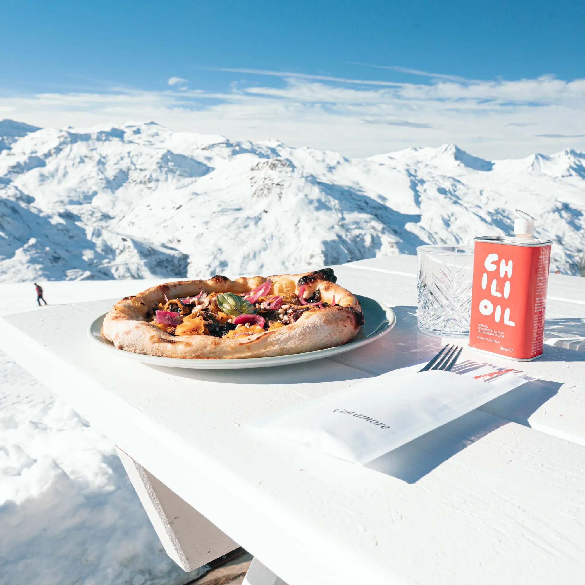 Pizza assiette sur une table blanche avec une canette d'huile au piment, un verre vide et des couverts enveloppés dans une serviette, en arrière-plan des montagnes enneigées sous un ciel bleu clair.