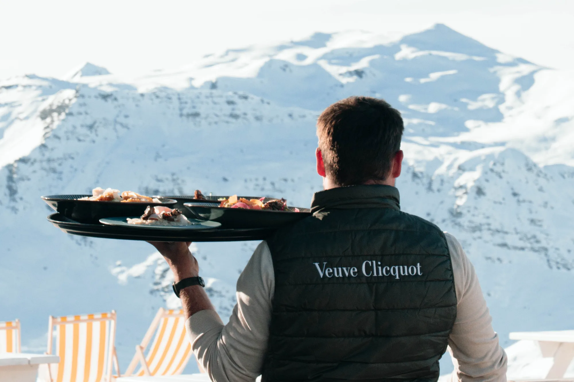Serveur vu de dos portant un gilet noir avec le texte Veuve Clicquot, tenant un plateau avec plusieurs assiettes de nourriture, devant un paysage de montagnes enneigées.