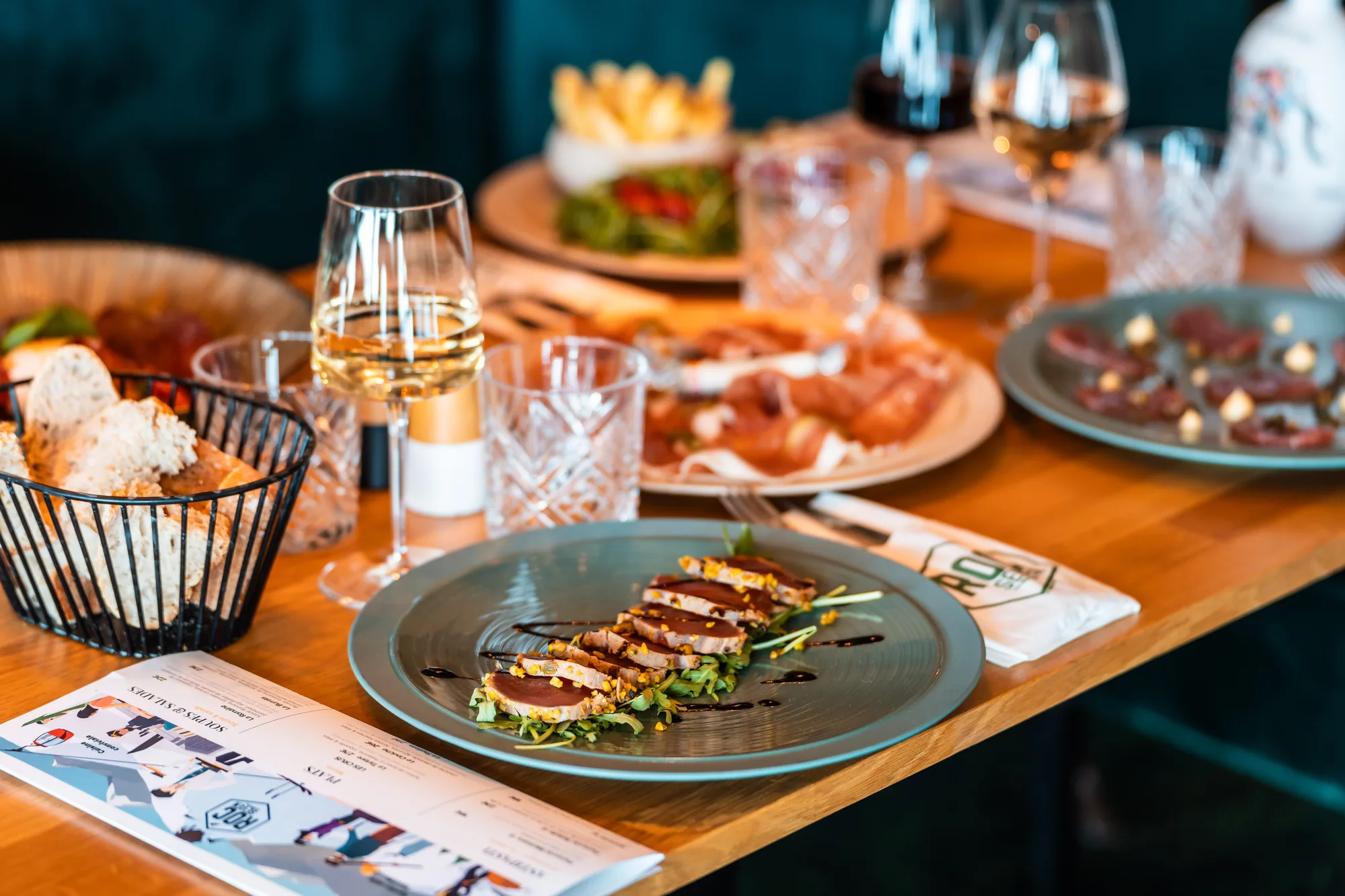 Table en bois avec assiettes de plats gastronomiques, un panier de pain, plusieurs verres à vin avec du vin blanc et rouge, et un menu ouvert au premier plan.