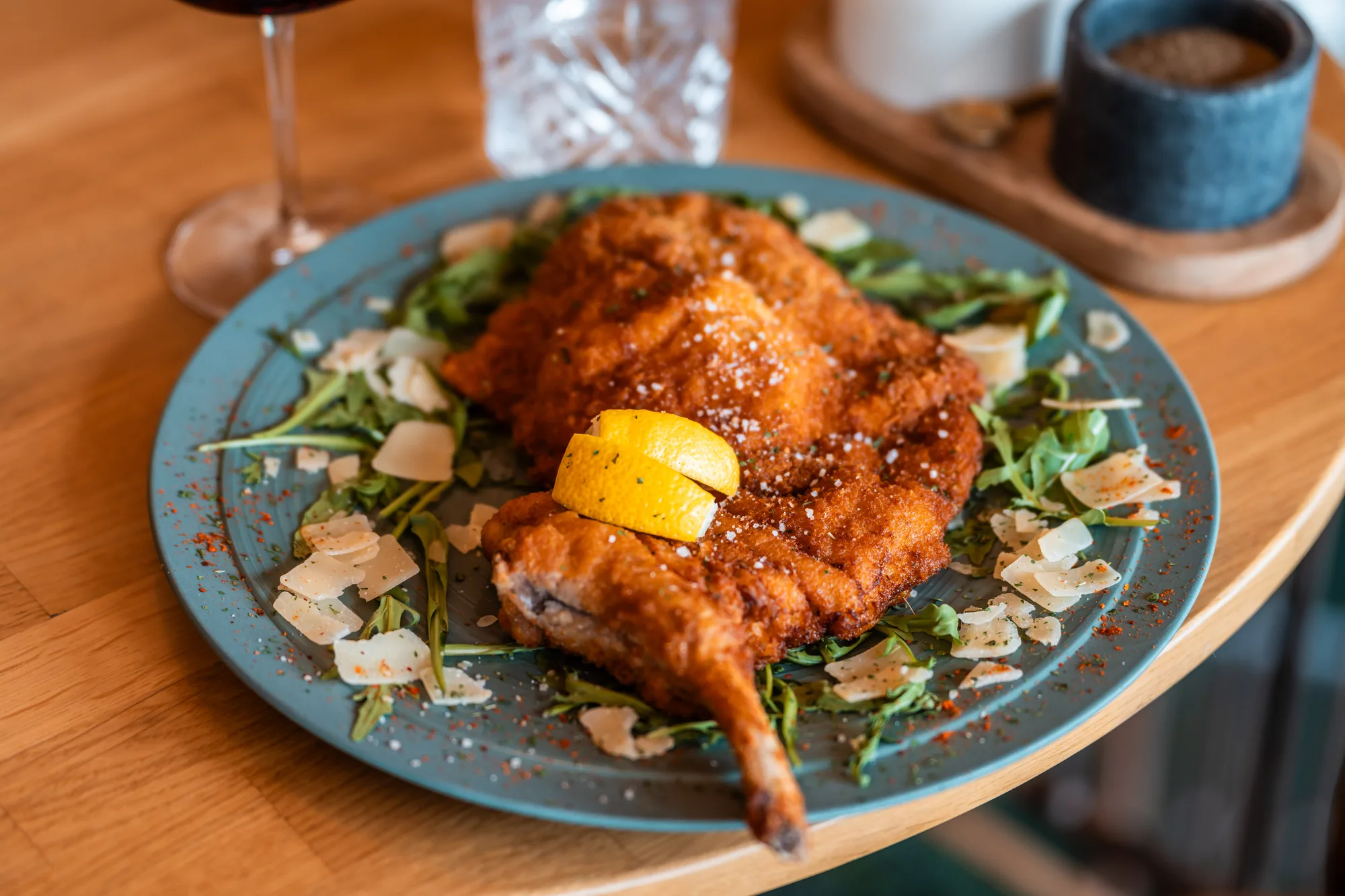 Assiette bleue avec un morceau de poulet pané croustillant sur un lit de roquette et de copeaux de parmesan, garni de quartiers de citron jaune, posé sur une table en bois.