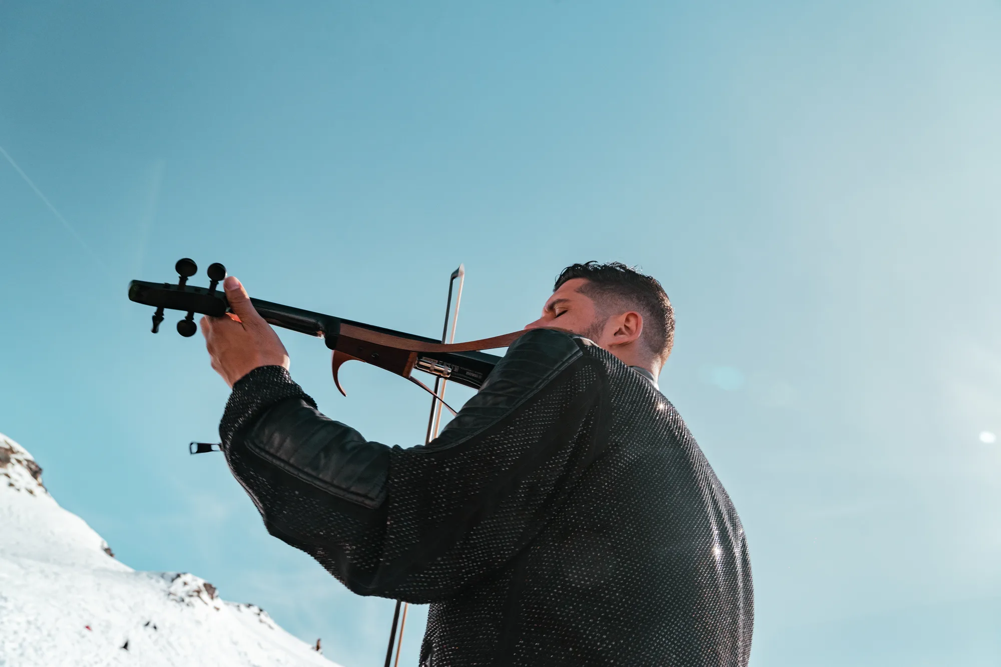 Un homme joue du violon électrique à l'extérieur sous un ciel bleu dégagé, avec une montagne enneigée en arrière-plan.