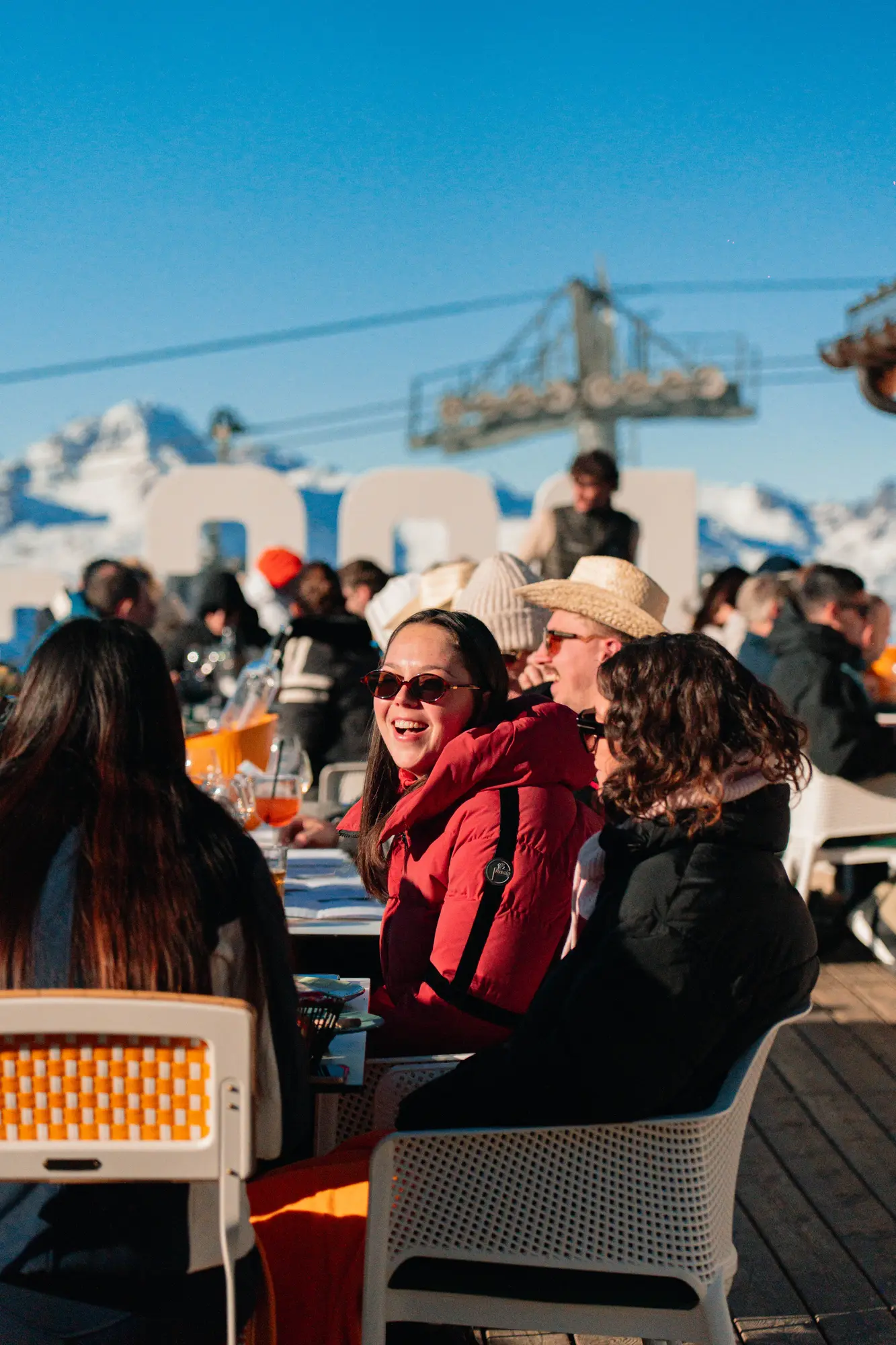 Groupe de personnes assises en terrasse sous un ciel bleu clair et entourées de montagnes enneigées, une femme souriante au centre porte une veste rouge et des lunettes de soleil.