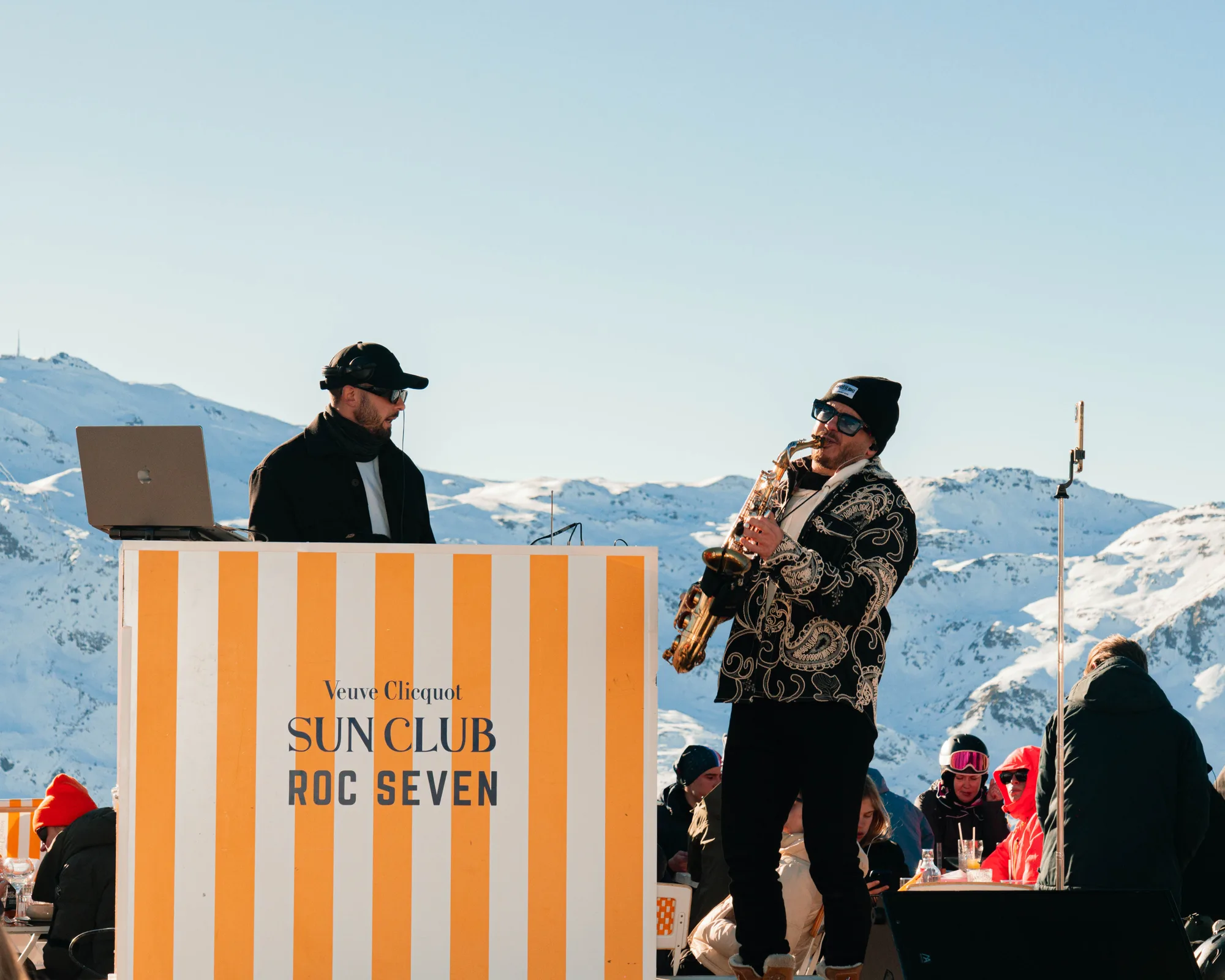 Un DJ portant une casquette noire et des écouteurs joue sur un ordinateur portable à côté d'un musicien jouant du saxophone, tous deux devant un pupitre rayé orange et blanc avec le texte « Veuve Clicquot Sun Club Roc Seven », avec des montagnes enneigées en arrière-plan et un groupe de personnes assises et debout au soleil.