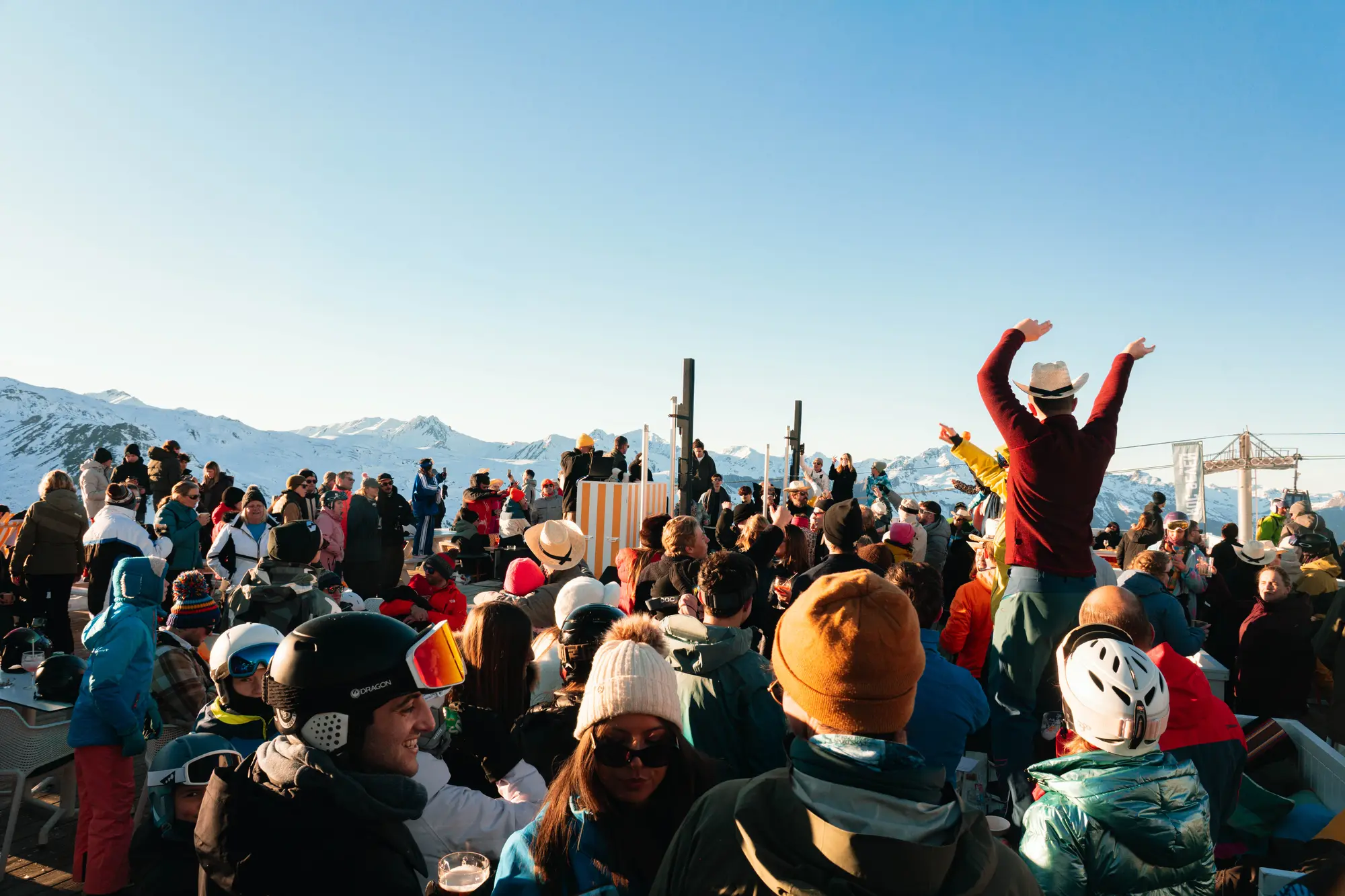 Un grand groupe de personnes vêtues de vêtements d'hiver se rassemble en plein air dans une station de montagne enneigée, avec des sommets enneigés en arrière-plan et un ciel clair.