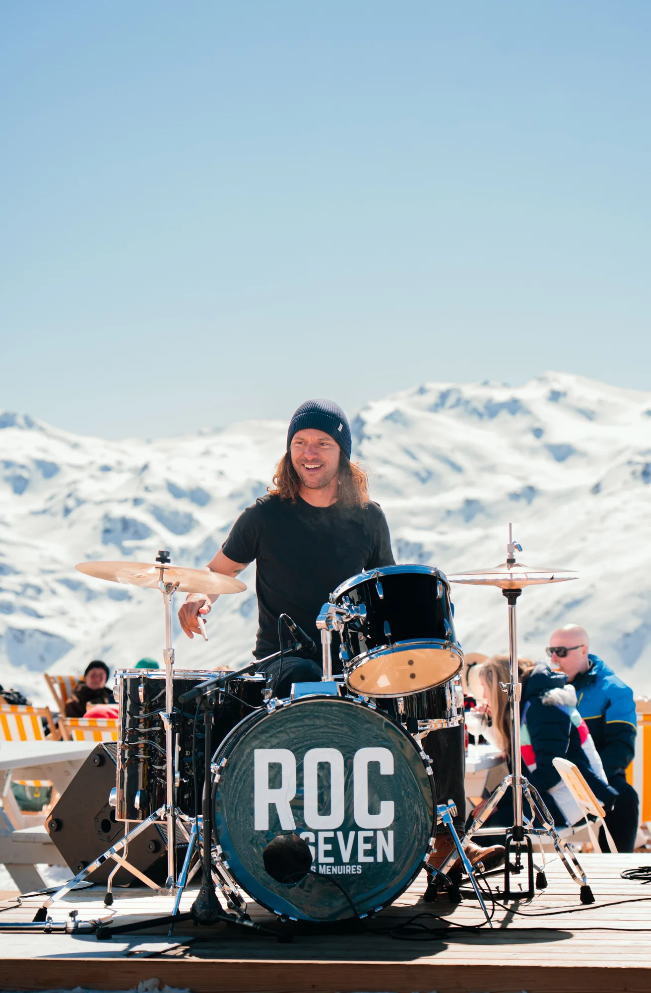 Un batteur souriant portant un bonnet noir joue d'une batterie avec le logo ROC SEVEN sur la grosse caisse, sur une scène extérieure avec des montagnes enneigées en arrière-plan et des personnes assises à des tables blanches.