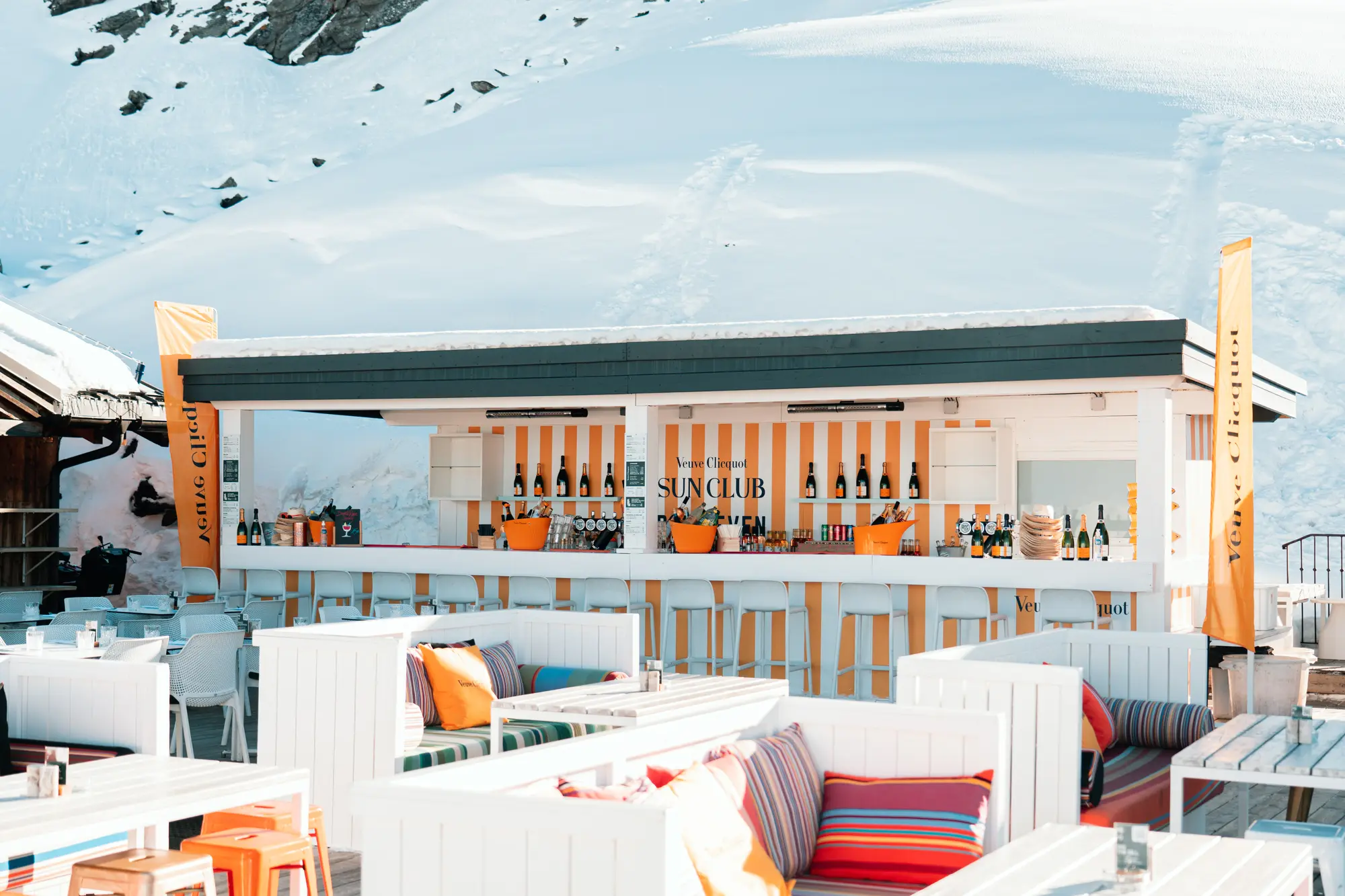 Bar extérieur blanc avec tabourets et canapés garnis de coussins colorés, situé dans un cadre enneigé avec montagnes en arrière-plan, décoré aux couleurs jaune et blanc de Veuve Clicquot, avec bouteilles de champagne visibles sur le comptoir.