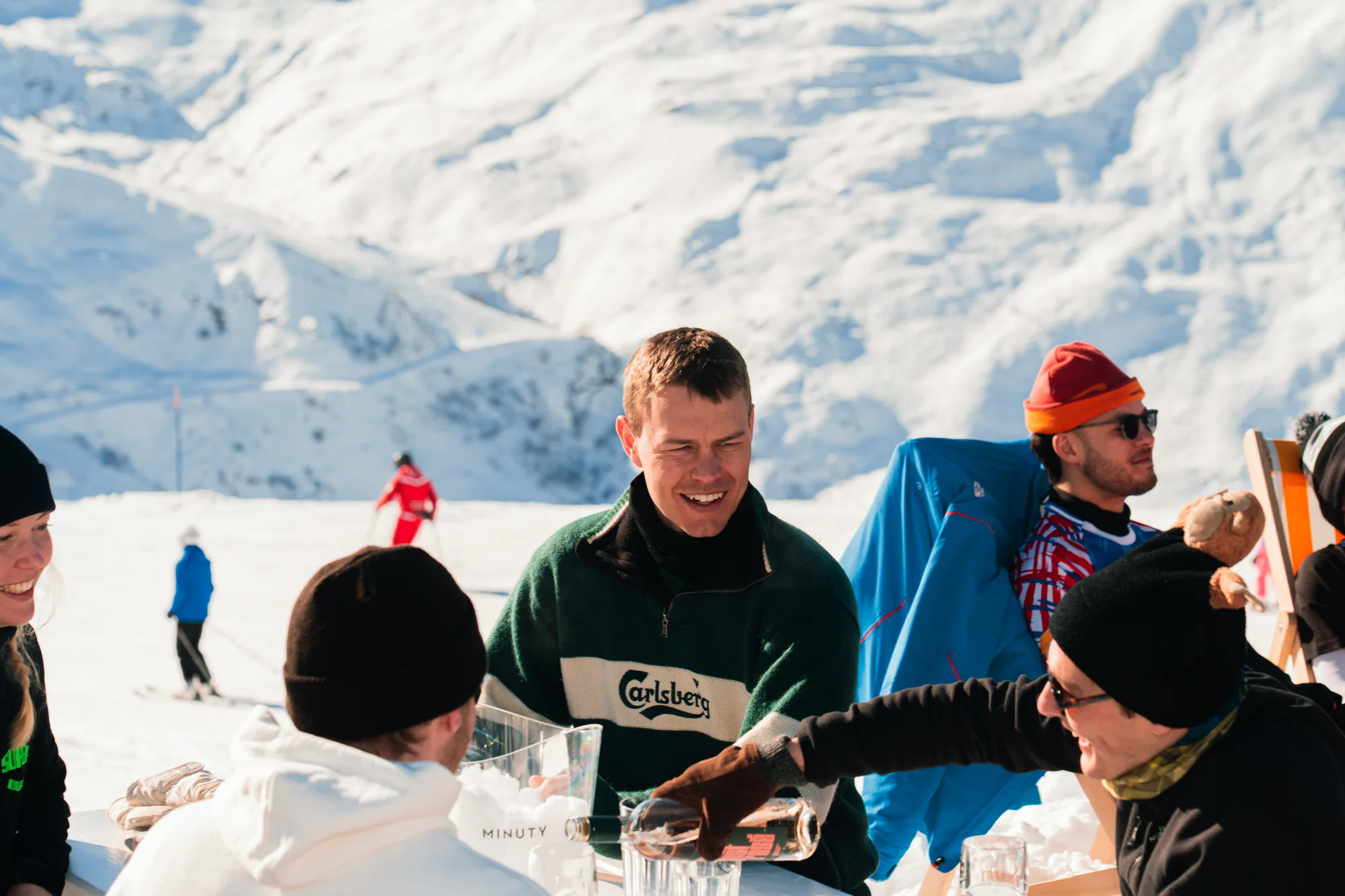 Un groupe d'amis souriants assis autour d'une table en plein air sur la neige, en montagne, avec des personnes en tenue de ski en arrière-plan et des montagnes enneigées.