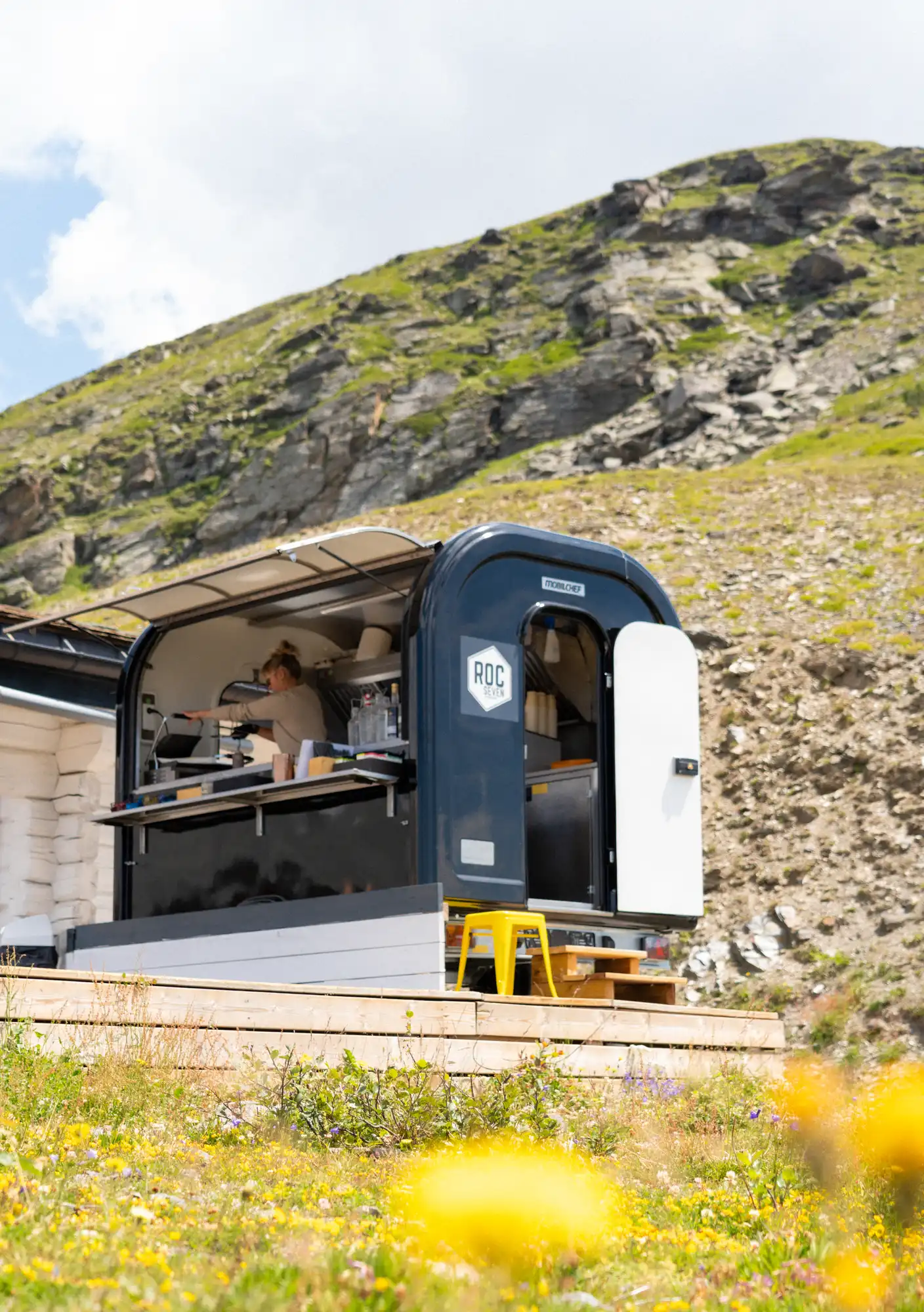 Une personne tenant un café dans une petite cuisine mobile noire avec un toit ouvert, installée sur une terrasse en bois dans un champ de fleurs au pied d'une colline rocheuse verte.