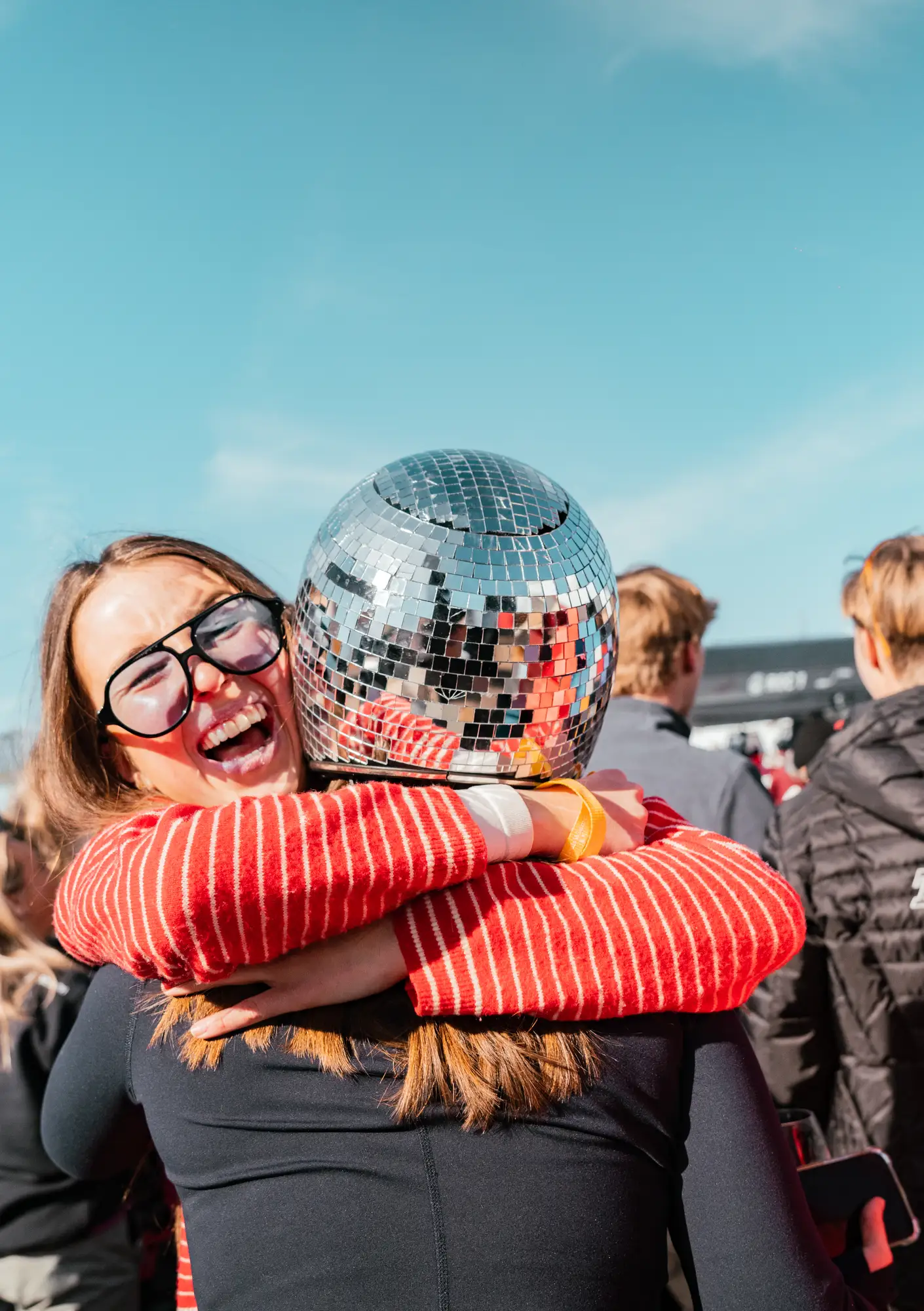 Une femme souriante portant de grandes lunettes noires serre dans ses bras une autre personne qui porte un casque en forme de boule à facettes lors d'un événement en plein air sous un ciel bleu.