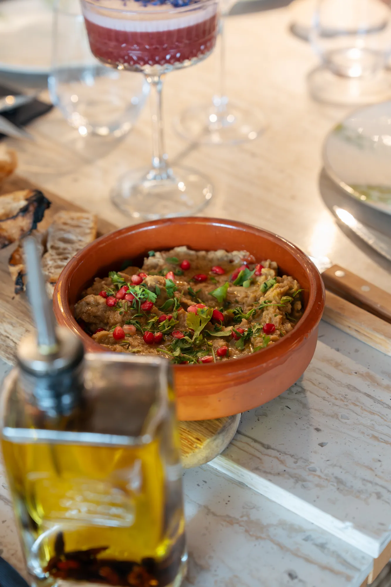 Bol en terre cuite contenant une purée à base d'aubergines garnie de graines de grenade et de coriandre fraîche, posé sur une planche en bois avec du pain grillé sur le côté, une bouteille d'huile d'olive floue au premier plan et un verre de boisson rouge en arrière-plan sur une table.