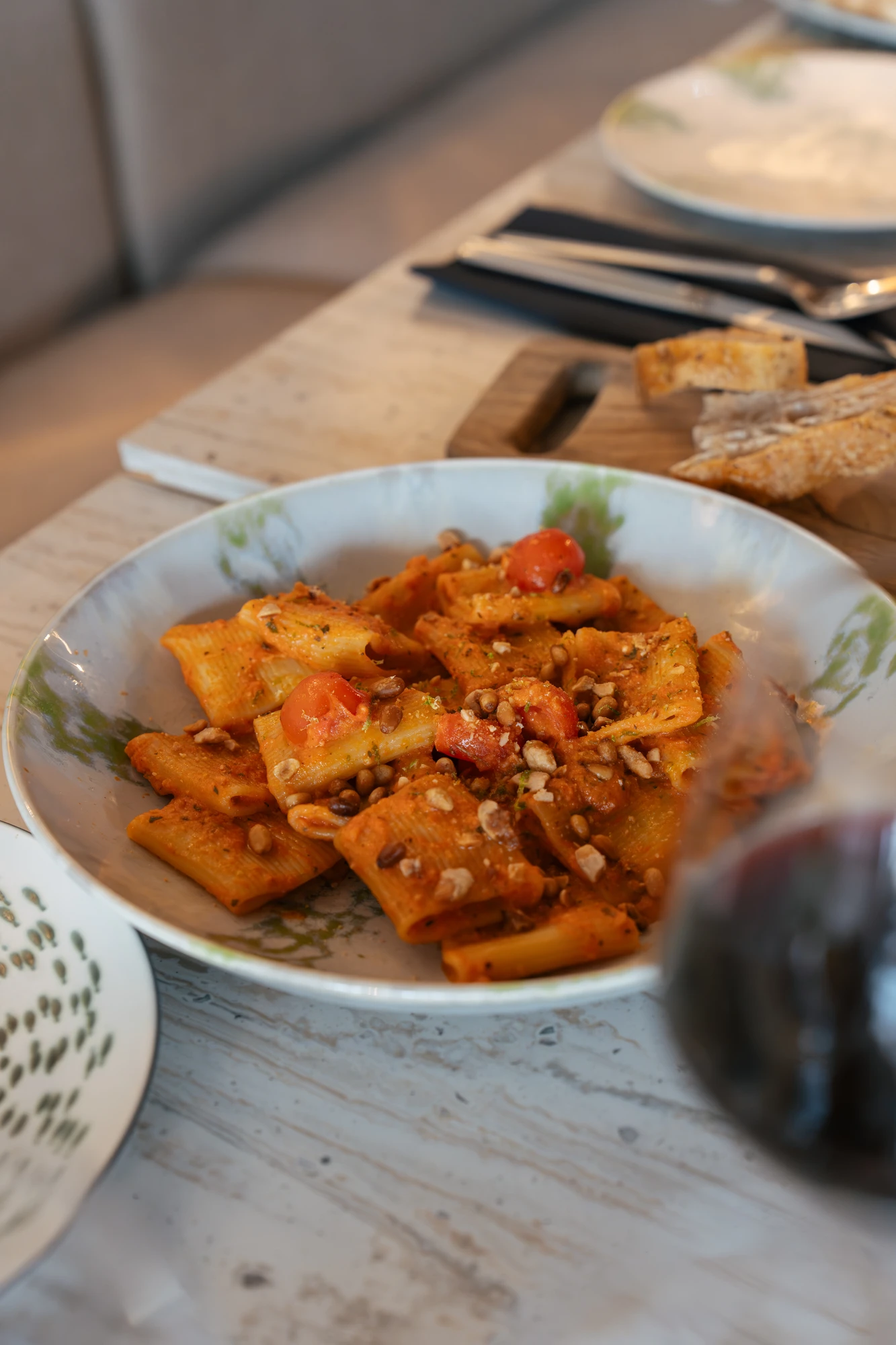 Assiette de pâtes rigatoni avec sauce tomate, tomates cerises et pignons de pin, posée sur une table avec un verre de vin rouge en avant-plan et du pain en arrière-plan.