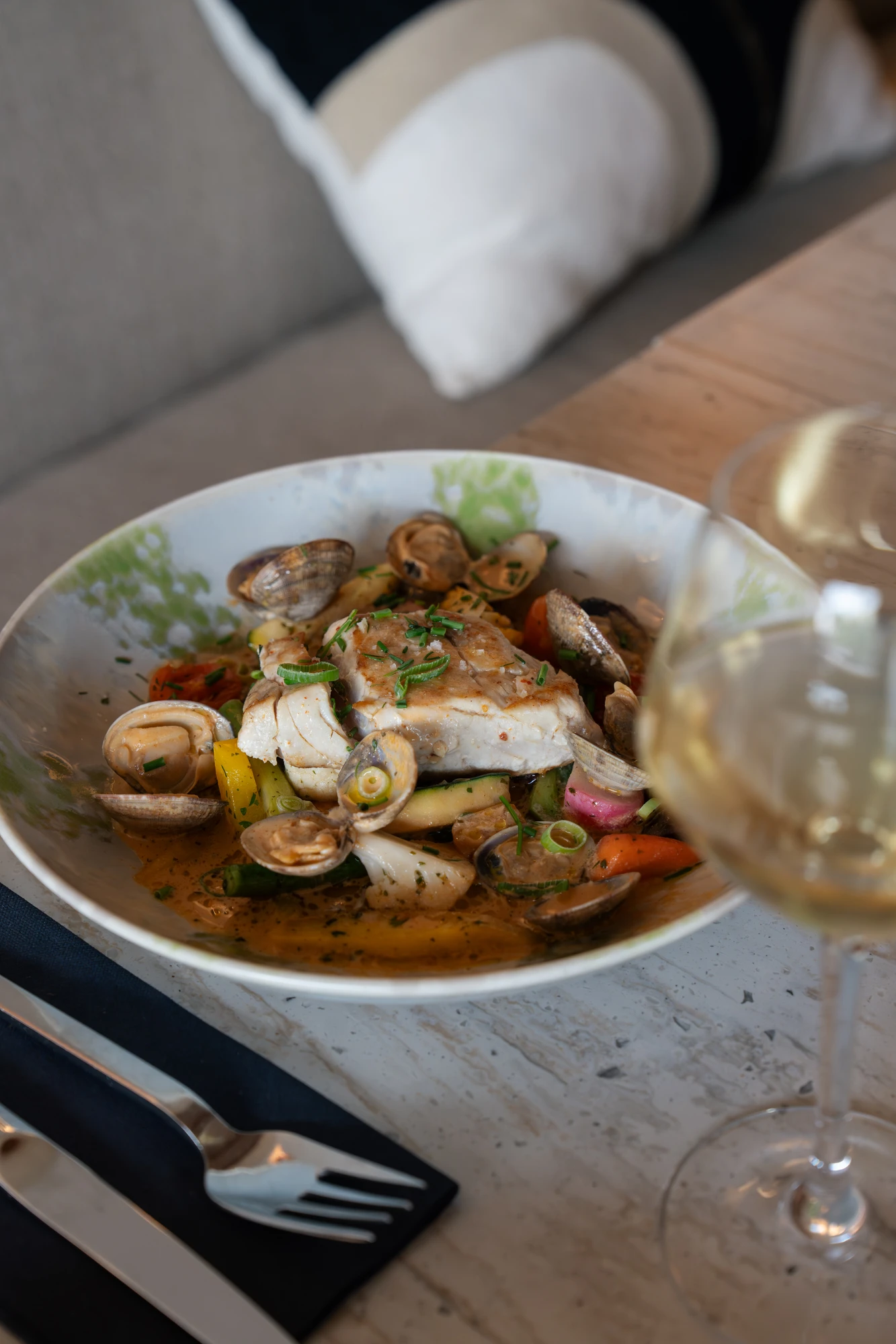 Assiette contenant un plat de poisson blanc poêlé accompagné de palourdes, légumes variés et herbes fraîches, posé sur une table avec un verre de vin blanc et des couverts sur une serviette noire.