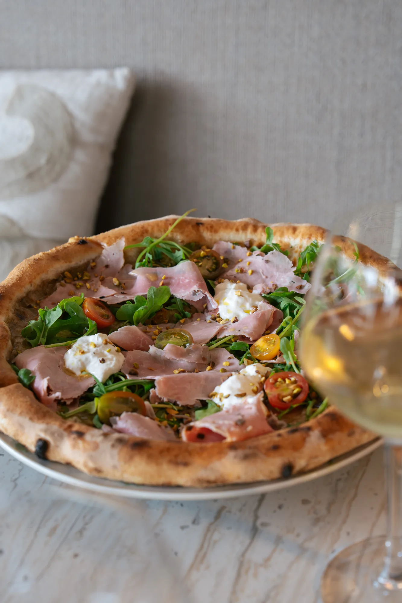 Pizza garnie de tranches de jambon, roquette, tomates cerises jaunes et rouges, morceaux de fromage frais et éclats de pistache, avec un verre de vin blanc flou au premier plan.