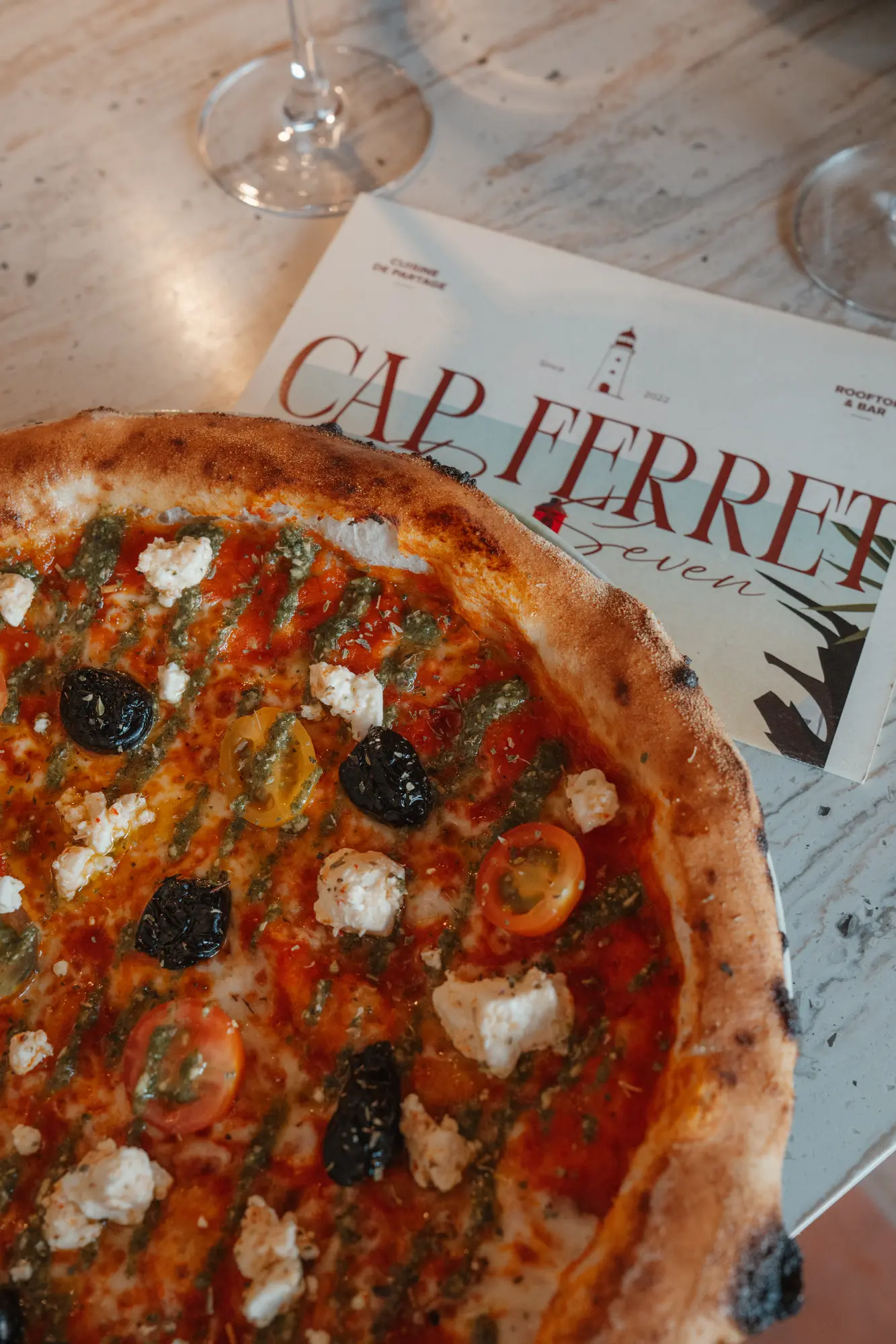 Pizza avec croûte dorée garnie de tomates cerises, olives noires, fromage blanc émietté et filet de pesto vert posée sur une table avec un menu marqué Cap Ferret en arrière-plan.