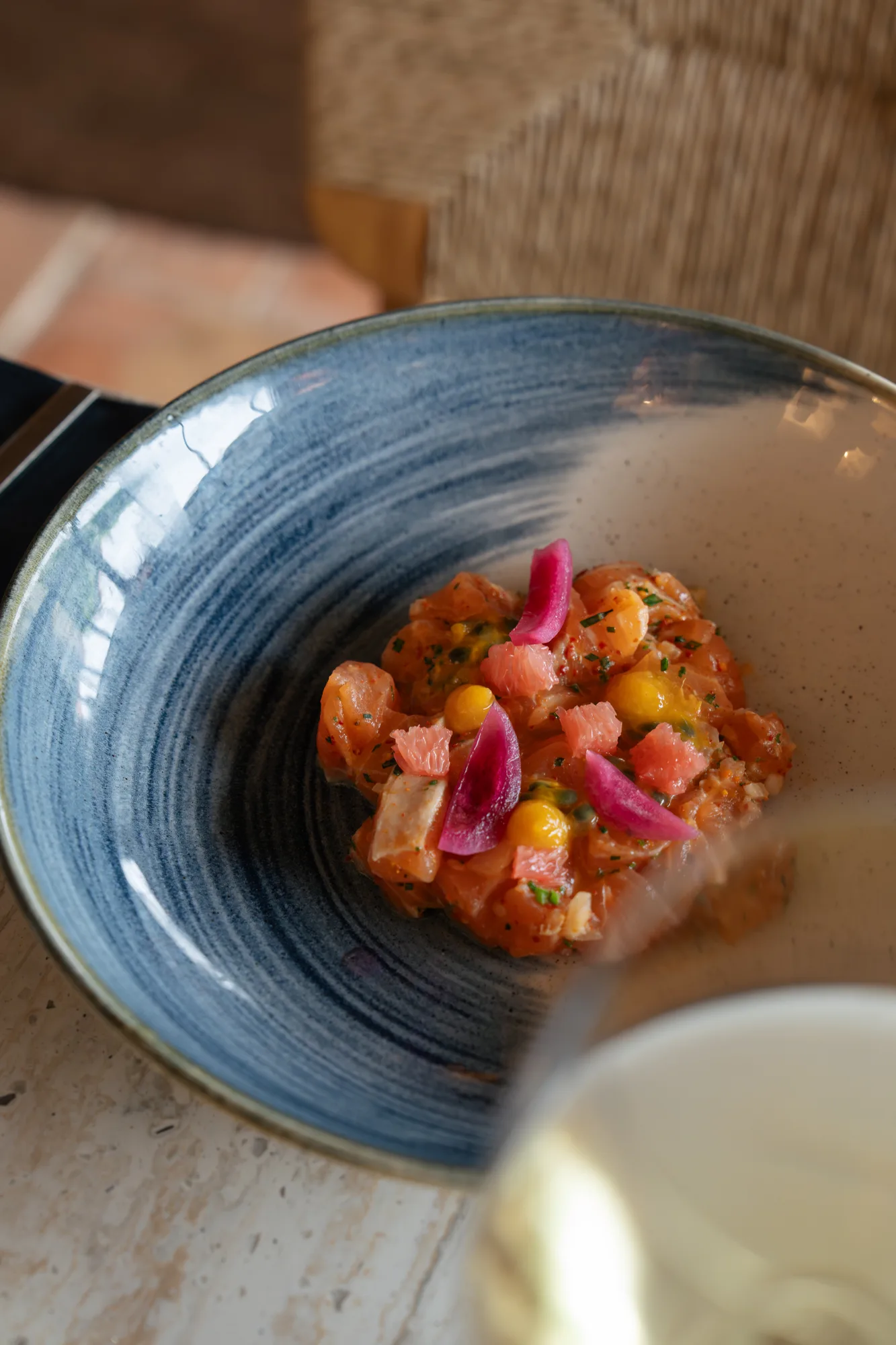 Assiette creuse avec un tartare de poisson cru décoré de quartiers de pamplemousse rose, de petites pousses vertes et de pétales de fleur violets, posée sur une table en pierre claire avec un verre flou en avant-plan.