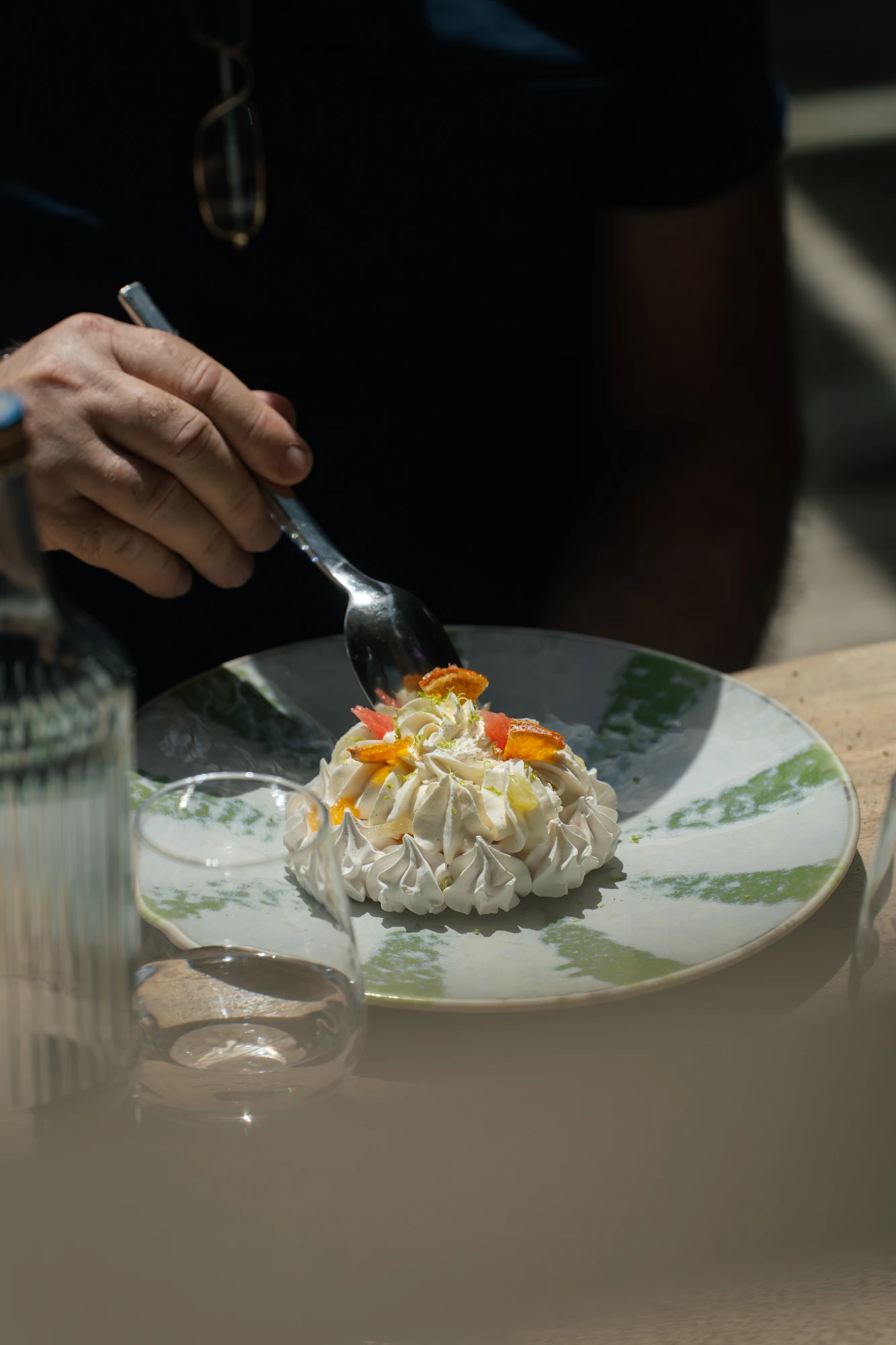 Une main tient une cuillère au-dessus d'un dessert élégant sur une assiette verte et blanche, garni de crème fouettée et de morceaux de fruits.