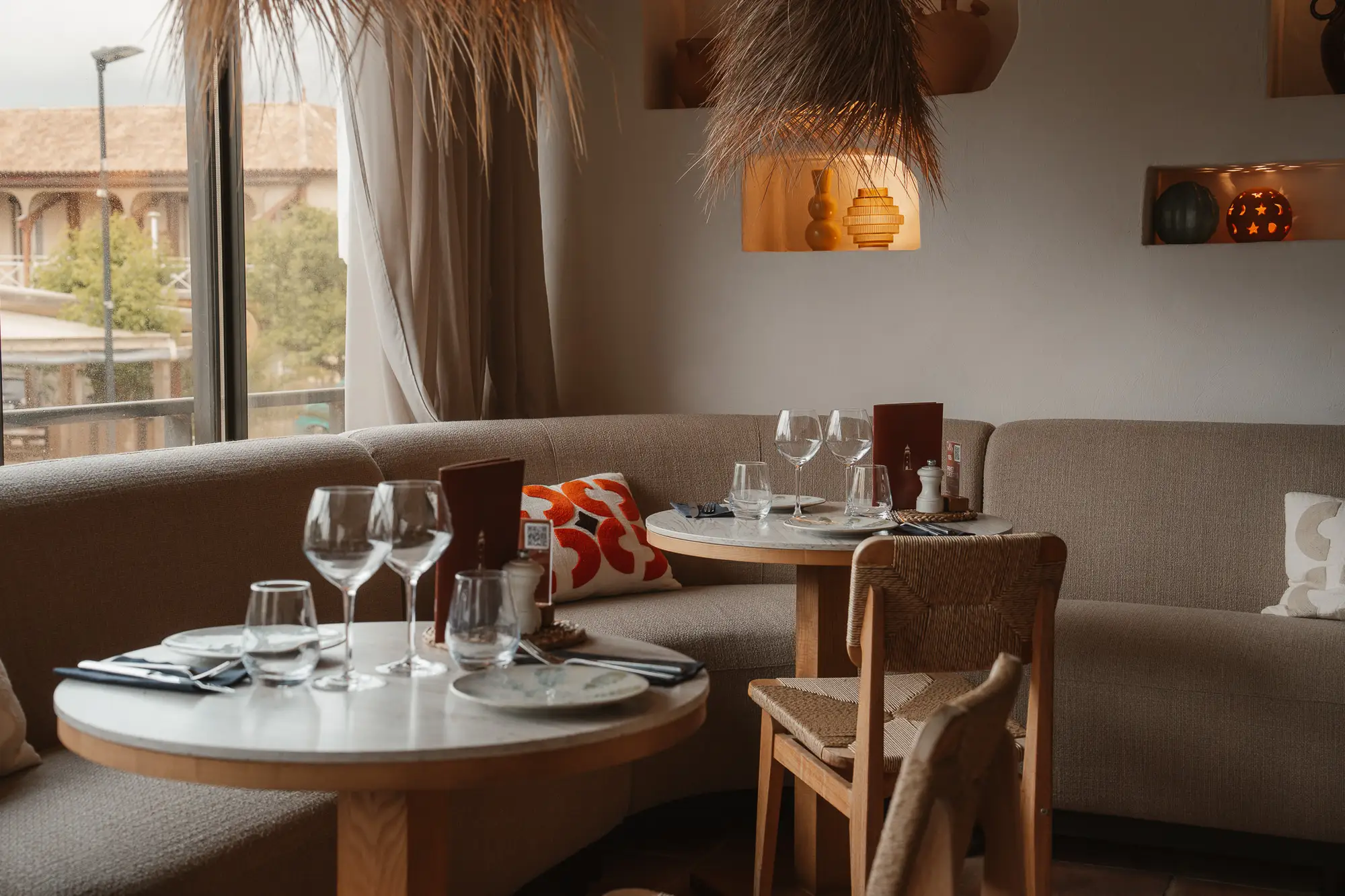 Coin repas chaleureux avec deux tables rondes en bois clair, nappes blanches, verres à vin et à eau, serviettes et couverts, banquettes beiges et coussins décoratifs, près d'une grande fenêtre.