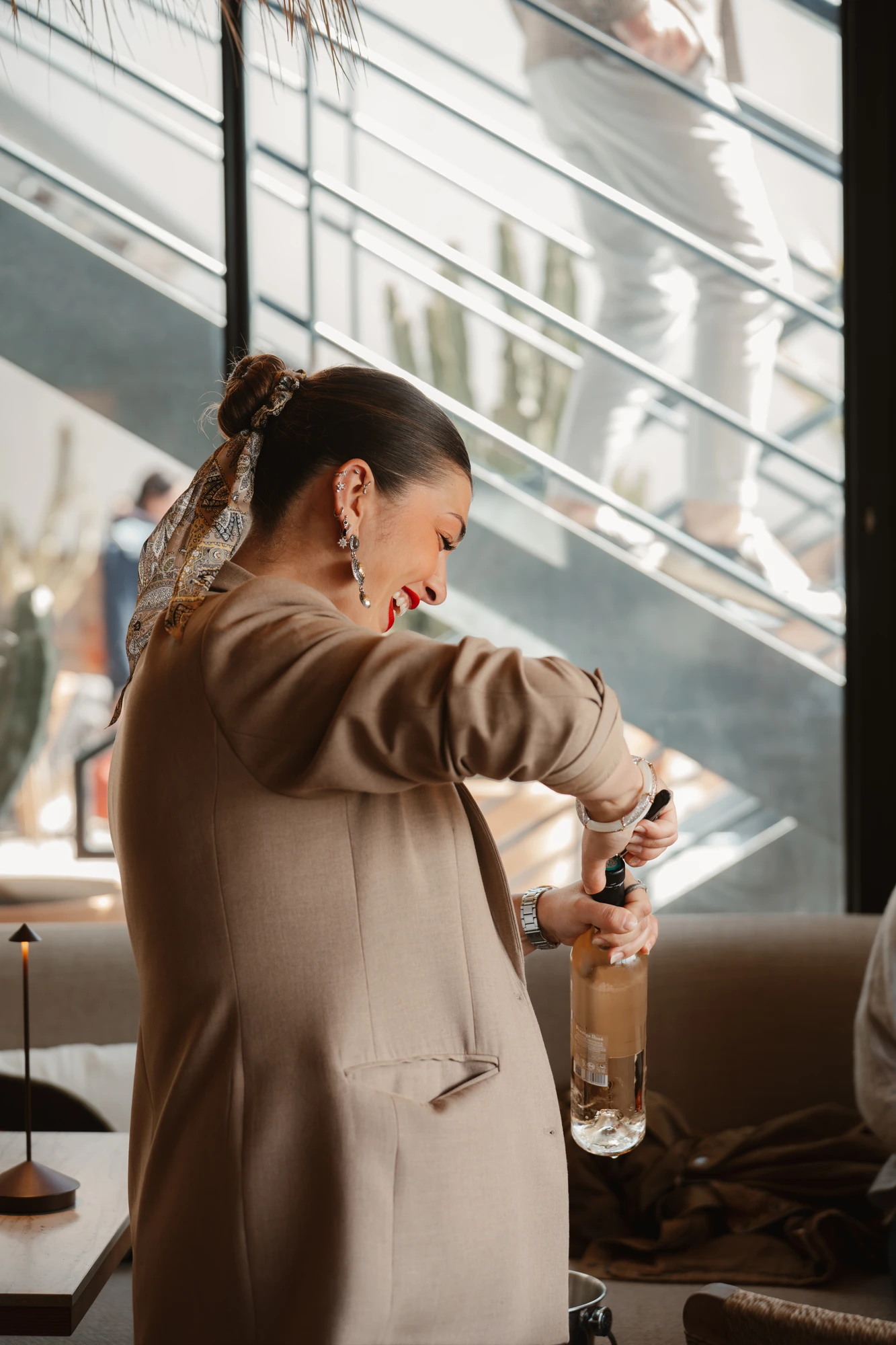 Femme souriante avec un chignon et un foulard dans les cheveux ouvre une bouteille de vin blanc à l'intérieur d'un café ou restaurant lumineux.