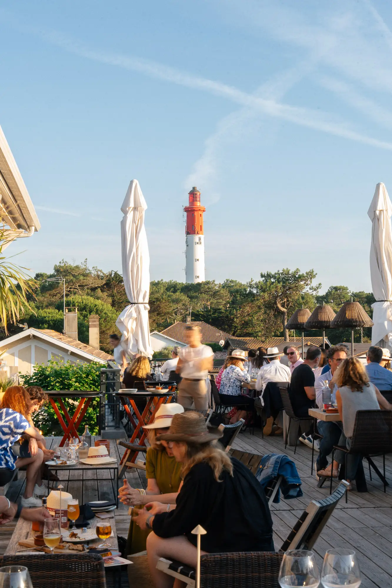 Terrasse extérieure d'un restaurant animée avec des personnes assises à des tables en bois et en osier, au centre un serveur en mouvement et en arrière-plan un phare blanc et rouge entouré de verdure sous ciel bleu.