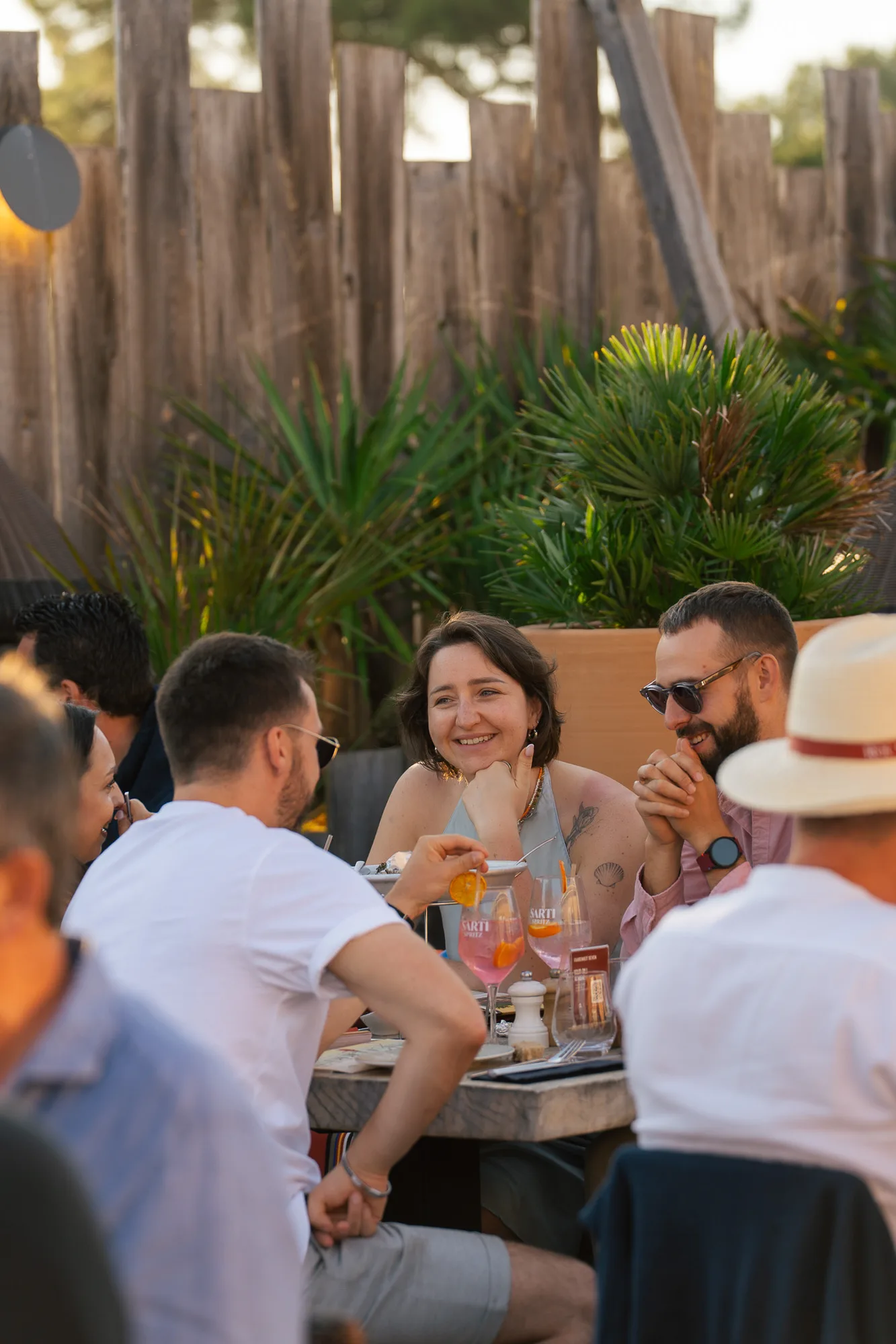 Un groupe de personnes assises autour d'une table en extérieur avec des boissons rosées décorées de tranches d'orange, discutant joyeusement dans une ambiance détendue avec des plantes et une clôture en bois en arrière-plan.
