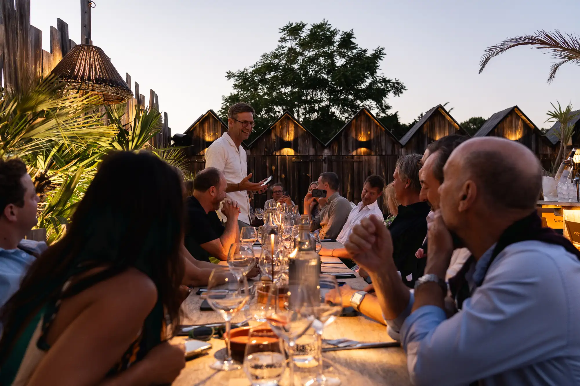 Groupe de personnes assises autour d'une longue table en bois lors d'un dîner en plein air au crépuscule, avec un homme debout qui parle et tient un téléphone, et des petites lampes éclairant subtilement la scène.