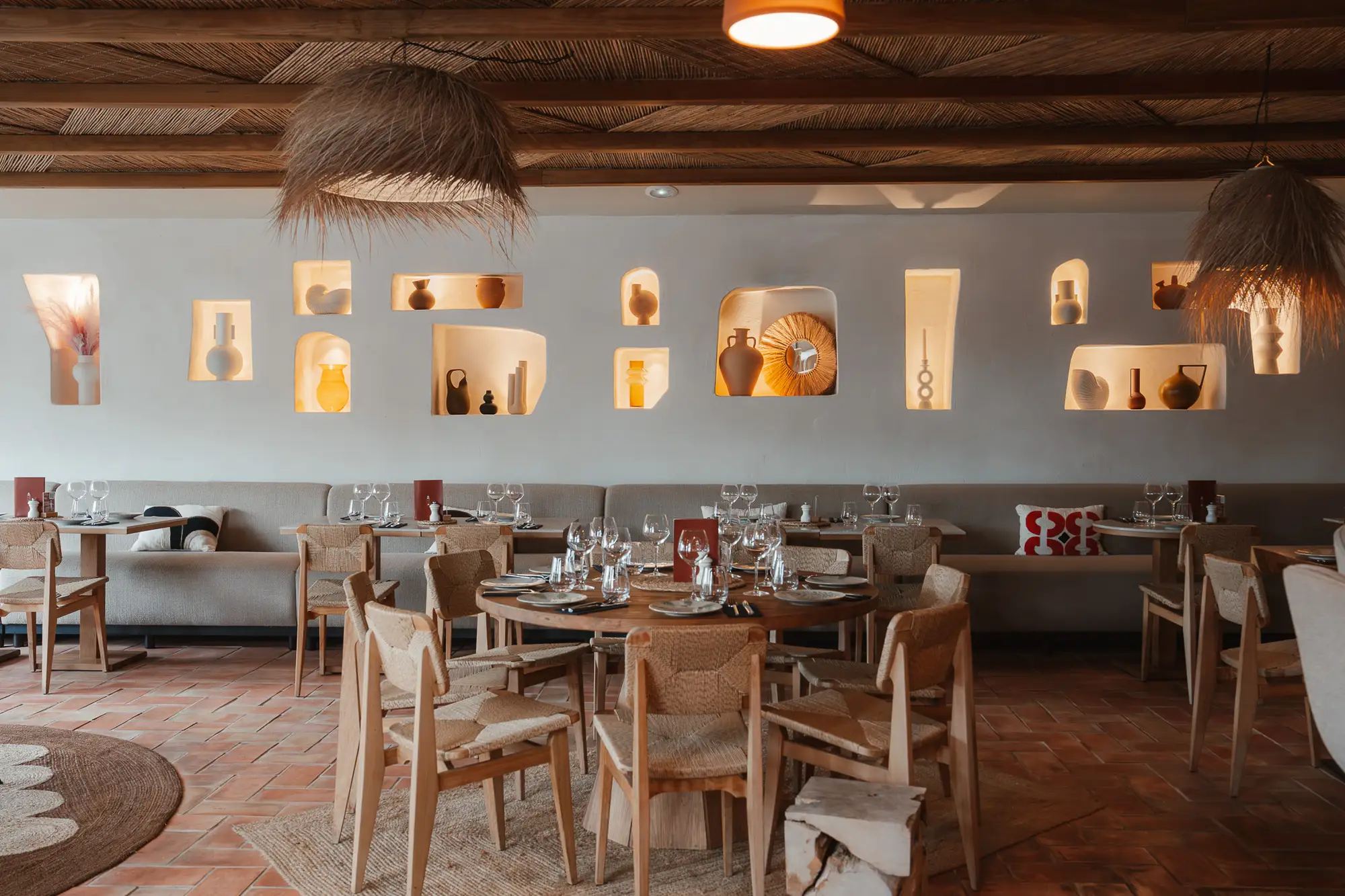 Intérieur d'un restaurant élégant avec des tables en bois rondes et carrées, chaises tissées, banquettes beiges, et une décoration murale avec des niches éclairées contenant des objets décoratifs en terre cuite et céramique, éclairage suspendu en fibres naturelles, et un plafond en bois avec des poutres apparentes.