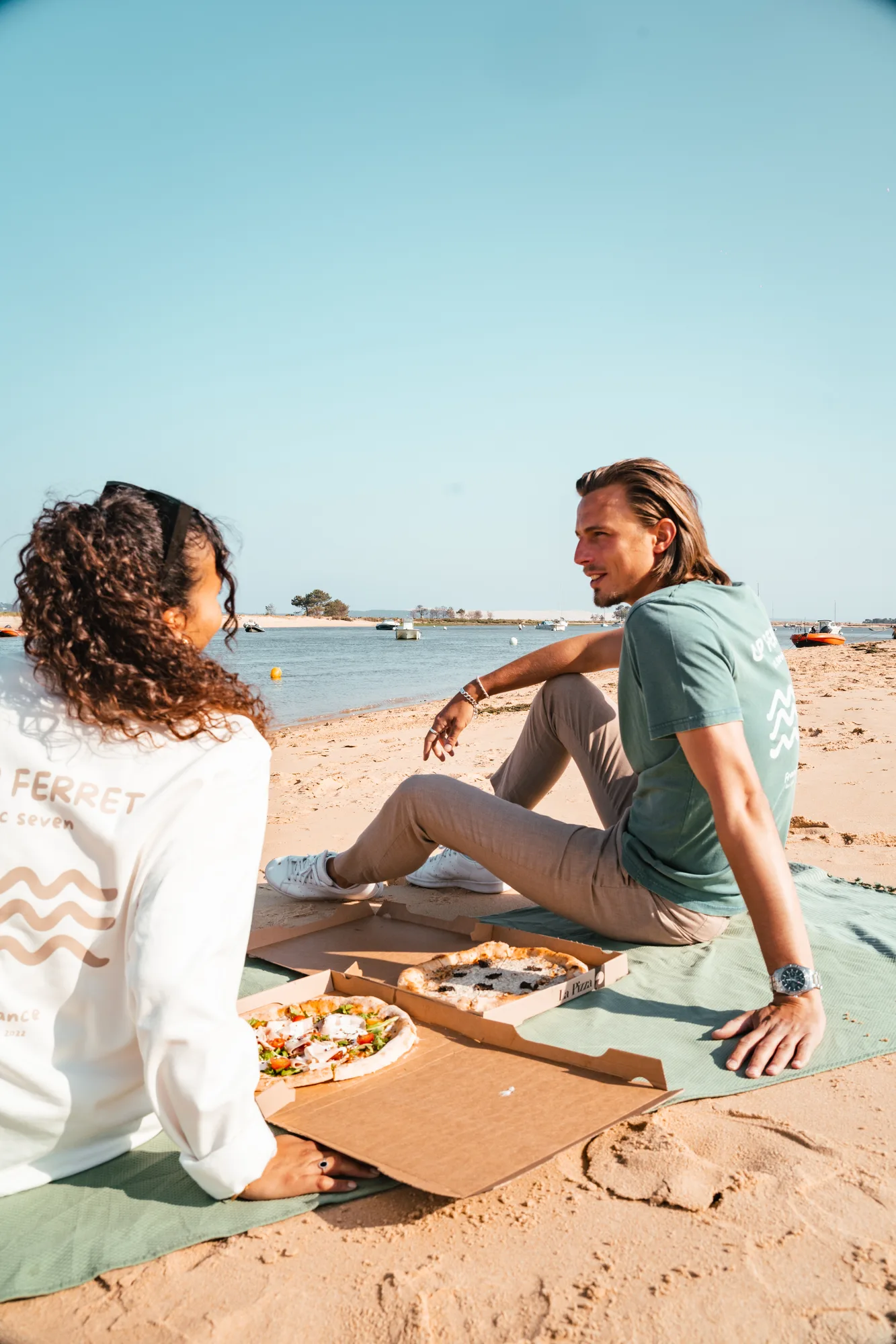 Un homme et une femme assis sur une plage sur une couverture verte, partageant deux pizzas dans des boîtes ouvertes, avec l'eau et des bateaux en arrière-plan sous un ciel bleu clair.