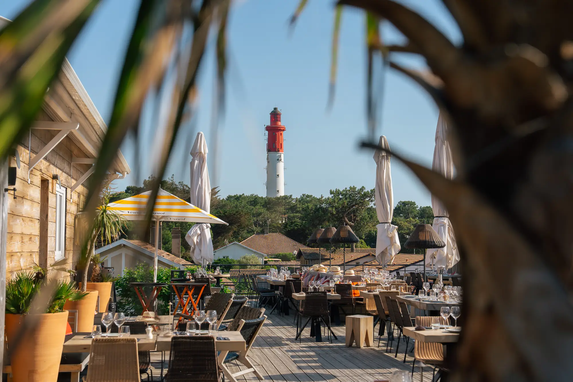 Terrasse en bois d'un restaurant avec plusieurs tables et chaises, dressées avec verres, vue sur un phare blanc et rouge au loin, ciel bleu clair et végétation verte autour.
