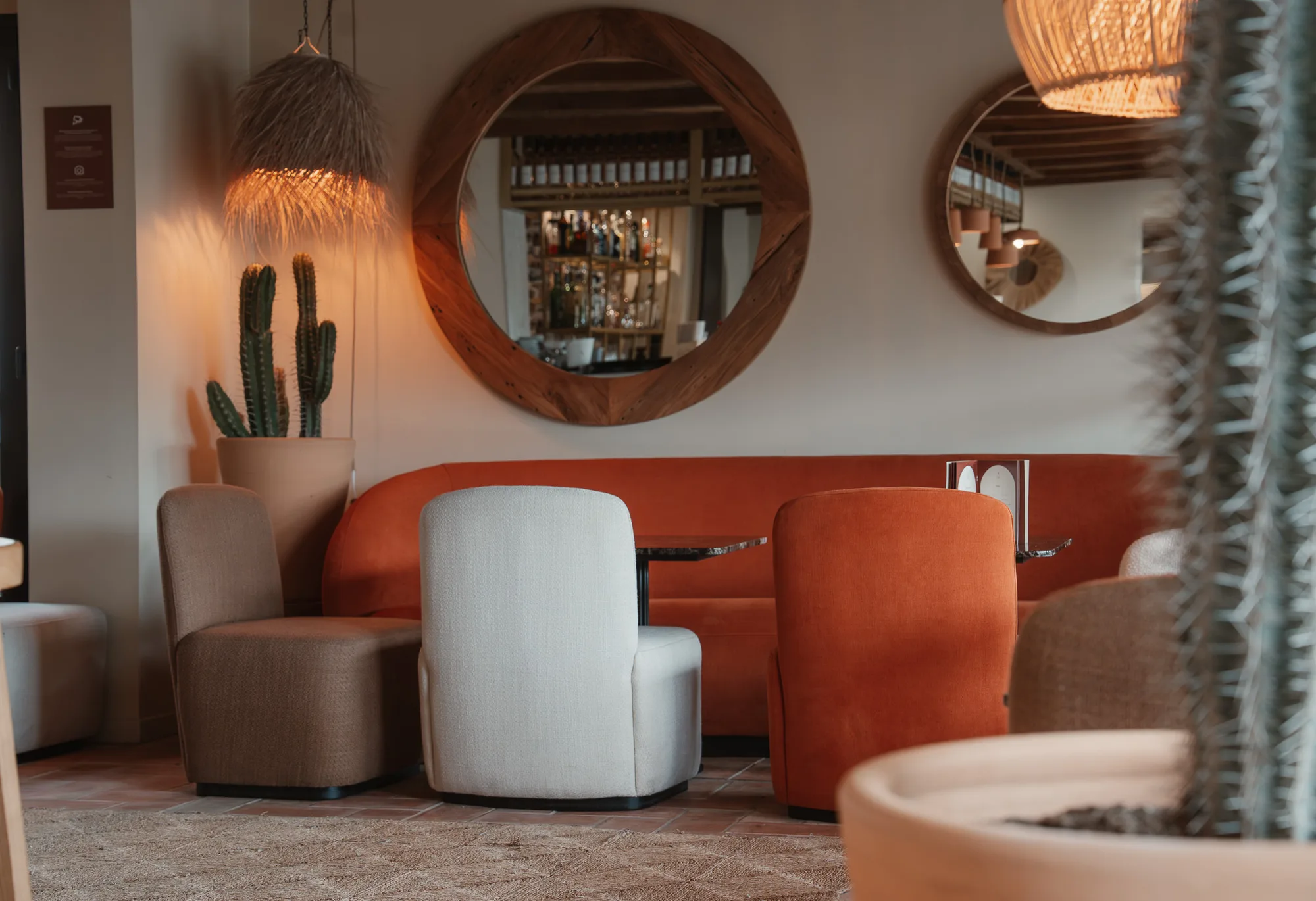 Coin salon chaleureux avec canapés orange, fauteuils beige et marron autour d'une petite table, cactus en pot, lampes suspendues et deux grands miroirs ronds sur un mur blanc.