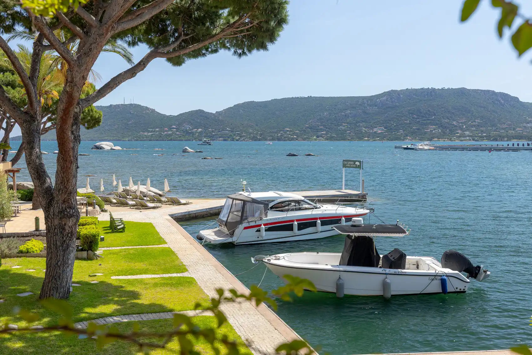 Vue sur un quai avec deux bateaux amarrés dans une eau calme, une pelouse verte avec des chaises longues et parasols fermés, et des collines verdoyantes en arrière-plan sous un ciel bleu clair.