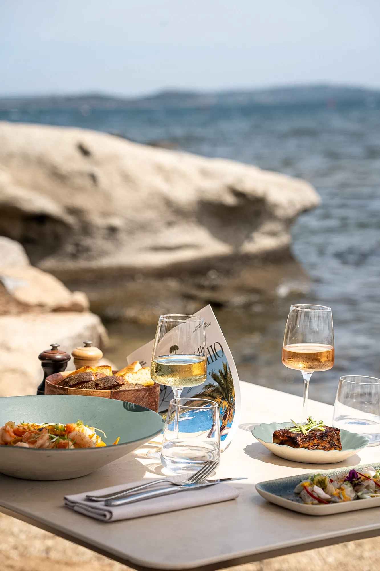 Table with an elegant seafood meal by the seaside, featuring a bowl of shrimp dish, a plate with grilled meat, a dish with a small salad, two glasses of white and rosé wine, water glasses, a basket of bread, salt and pepper shakers, and silverware on a napkin, with rocky shore and calm sea in the background.