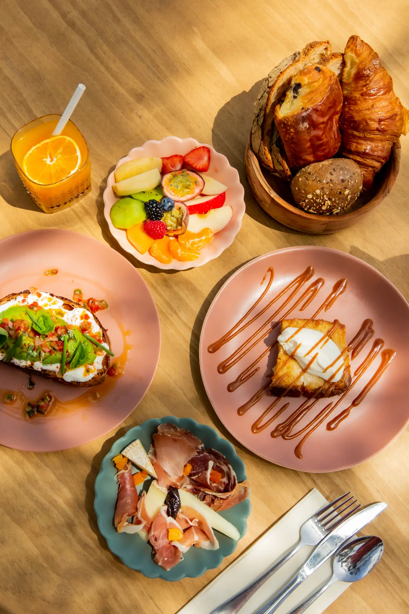 Un petit déjeuner comprenant un verre de jus d'orange avec une tranche d'orange, un bol de fruits frais variés, un panier de pains et viennoiseries, une assiette avec une tartine garnie de fromage frais, avocat et légumes, une assiette de charcuterie et fromages, et une assiette avec un dessert saupoudré de sauce caramel, accompagné de couverts sur une serviette.