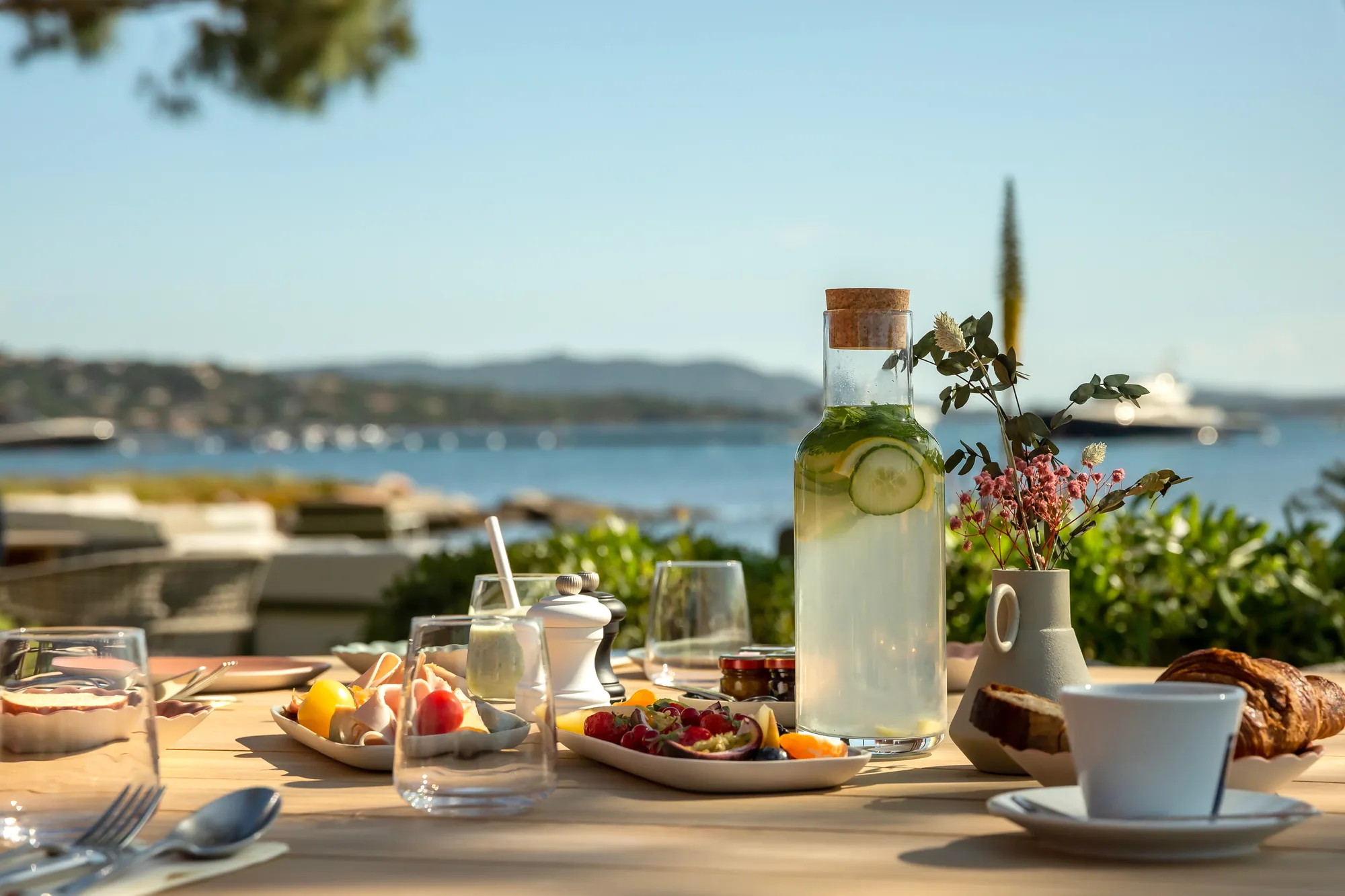 Table set for breakfast outdoors with a clear glass carafe of infused water with cucumber and lemon slices, plates of fruit and pastries, a cup and saucer, a small vase with pink flowers, and a sea view in the background under a clear sky.