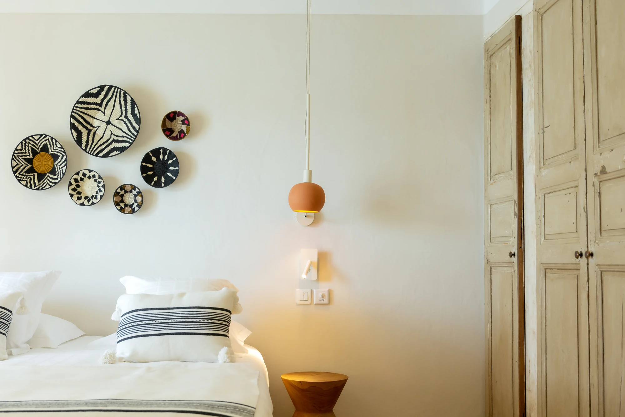 Chambre avec un lit double décoré de coussins blancs à rayures noires, sept paniers muraux décoratifs noirs et blancs accrochés au mur, une lampe suspendue orange et une table de chevet en bois clair près d'une armoire en bois patiné.
