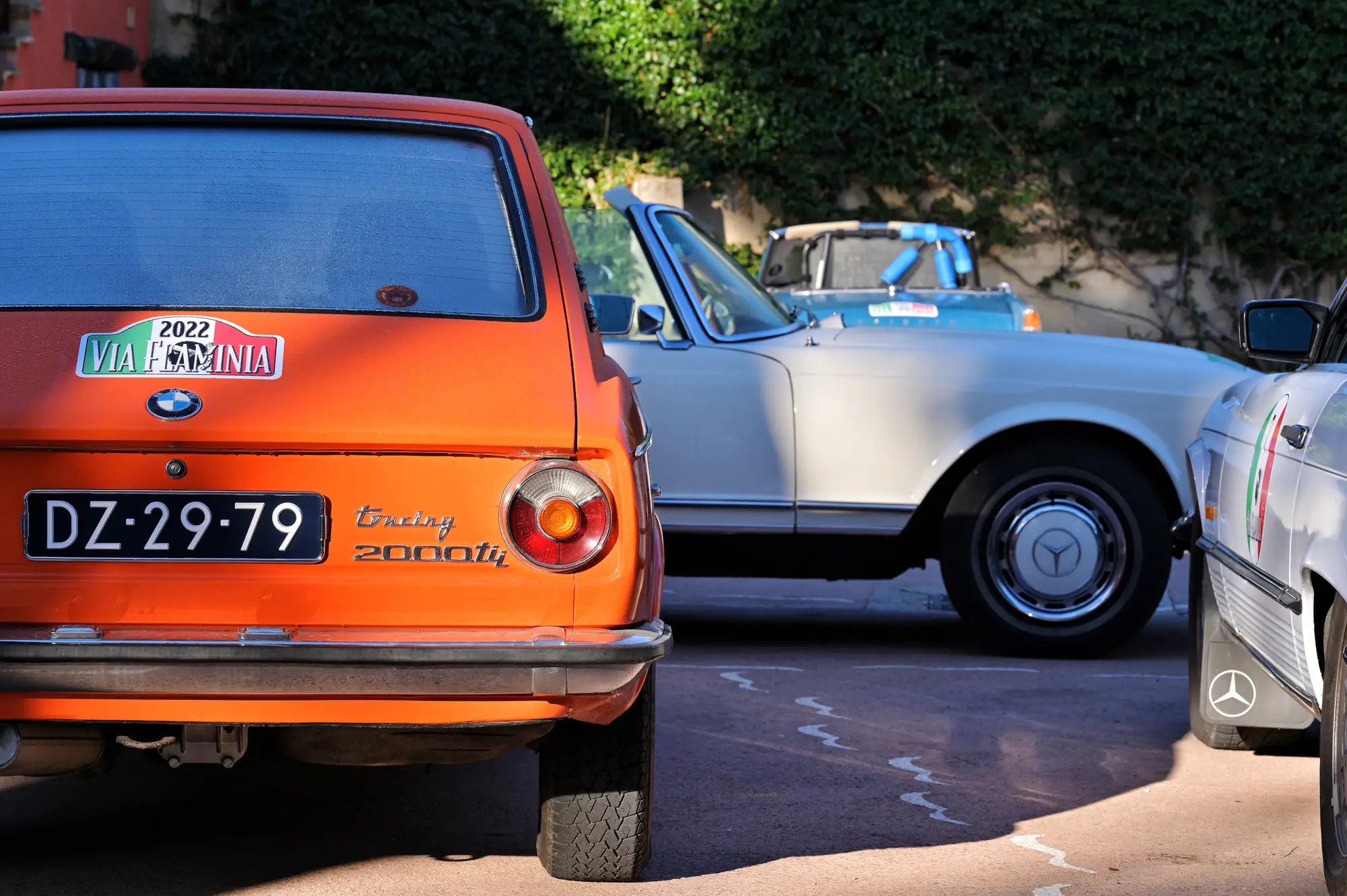 Vue arrière d'une BMW orange 2000 tii avec plaque d'immatriculation DZ-29-79, à côté d'une voiture blanche cabriolet Mercedes-Benz garée, partiellement visible, avec un décor de murs et de végétation en arrière-plan.