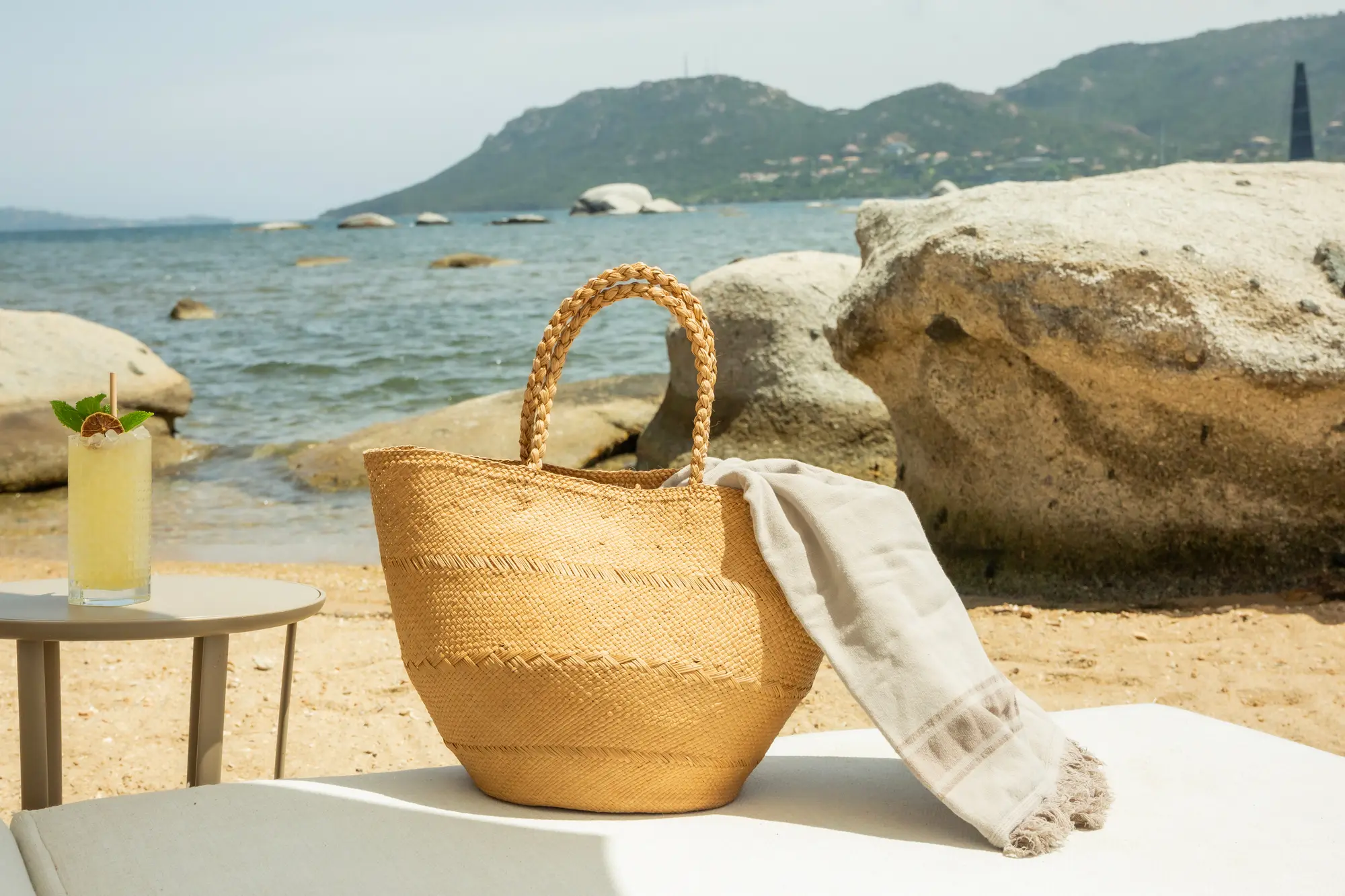 Sac de plage en paille clair avec une serviette beige posé sur une chaise longue près de la mer avec des rochers en arrière-plan, et un cocktail décoré de menthe sur une petite table ronde à côté.