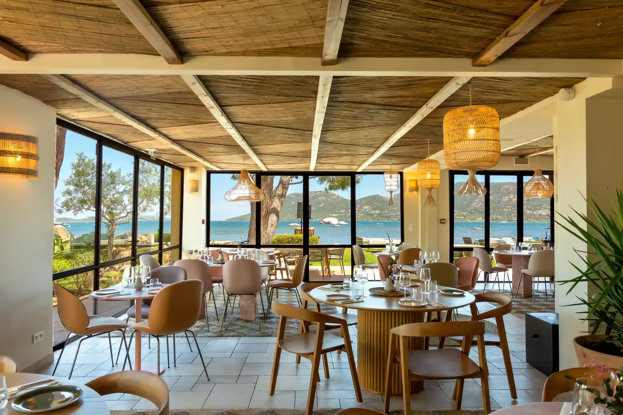 Intérieur d'un restaurant élégant avec des tables rondes et chaises modernes, des luminaires en osier suspendus, un plafond en bois, et de grandes fenêtres offrant une vue sur la mer et des collines au loin sous un ciel dégagé.