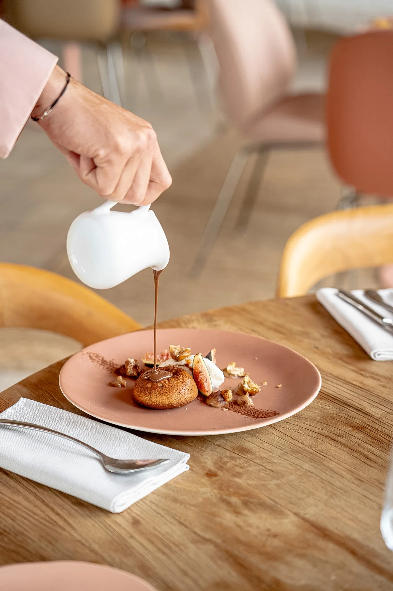 Une main versant du chocolat fondu d'une petite cruche blanche sur un dessert rond servi sur une assiette rose, accompagné de morceaux de fruits et noix, posé sur une table en bois avec une serviette blanche et une cuillère.