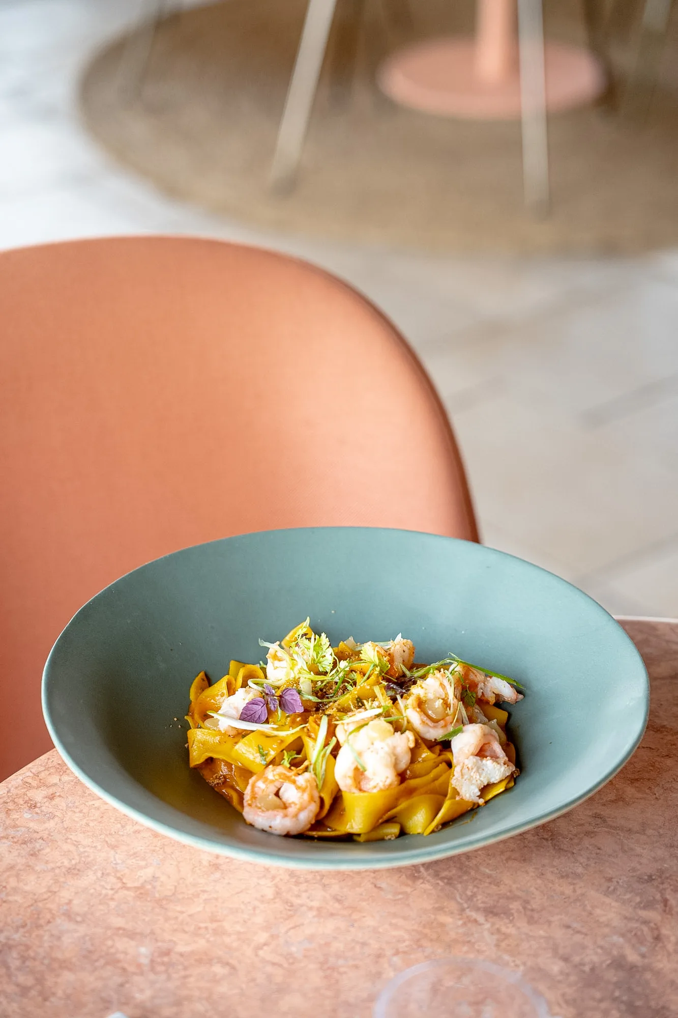 Plat de pâtes larges jaunes servies avec des crevettes garnies d'herbes fraîches dans un bol vert, posé sur une table en pierre rose avec une chaise orange en arrière-plan.