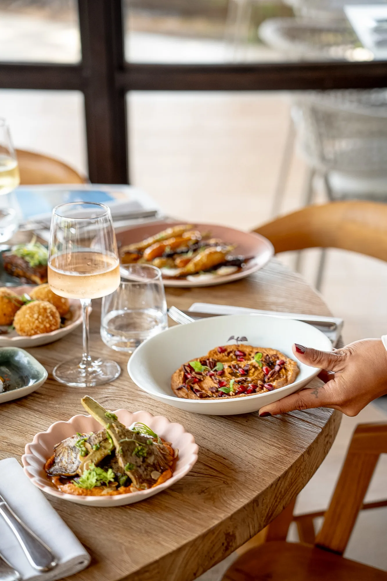 Une table en bois avec plusieurs plats gastronomiques disposés, dont un bol blanc avec une purée garnie de graines rouges et d'herbes, tenu par une main, un verre de vin rosé, un verre d'eau, et un autre plat avec des artichauts assaisonnés.