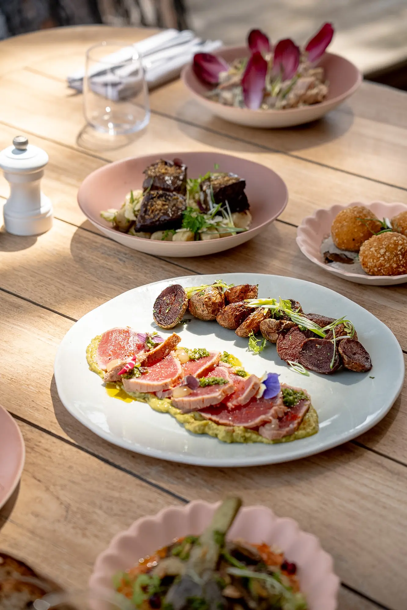 Assiette blanche avec des tranches de viande rosée sur une purée verte, accompagnées de petites pommes de terre rôties et de chips de légumes garnies de fines herbes, sur une table en bois avec plusieurs autres plats en arrière-plan.