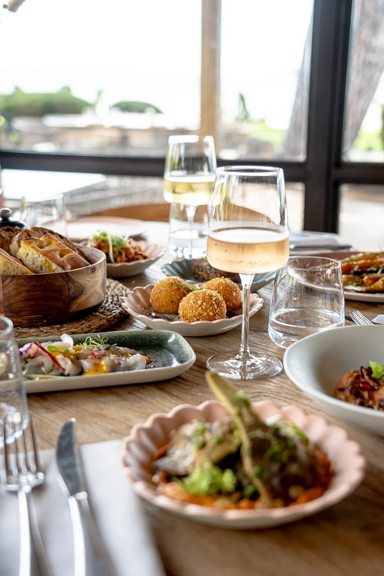 Table dressée avec plusieurs plats gastronomiques dont des croquettes, du pain, et des assiettes avec diverses préparations, accompagnés de verres de vin et d'eau, dans un décor lumineux avec une grande fenêtre en arrière-plan.