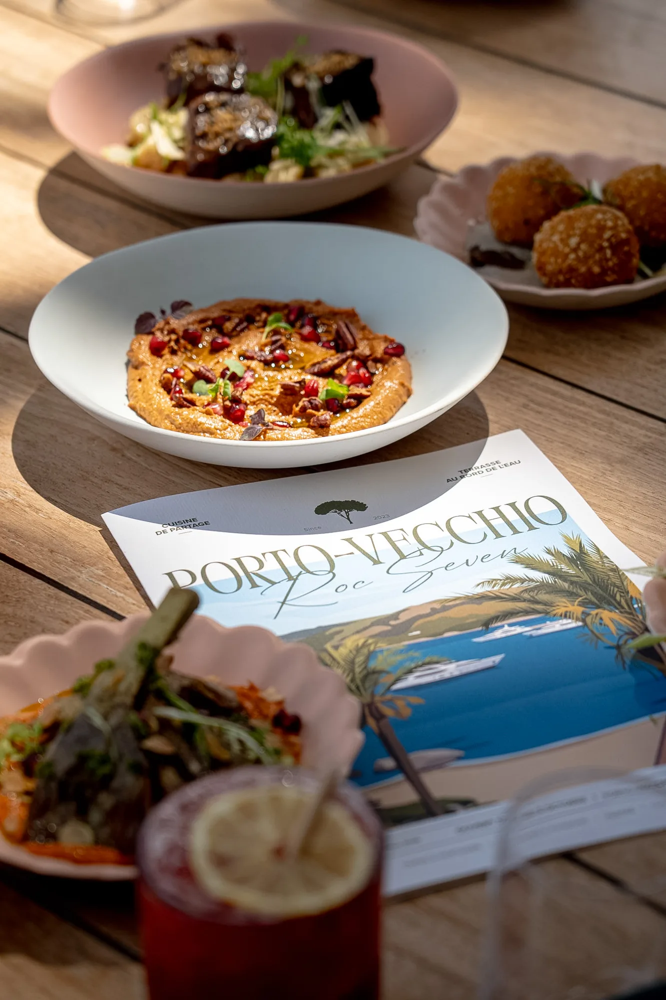 Table en bois avec plusieurs plats gastronomiques et un menu de restaurant intitulé Porto-Vecchio Roc Seven avec une illustration de yachts sur l'eau et des palmiers.