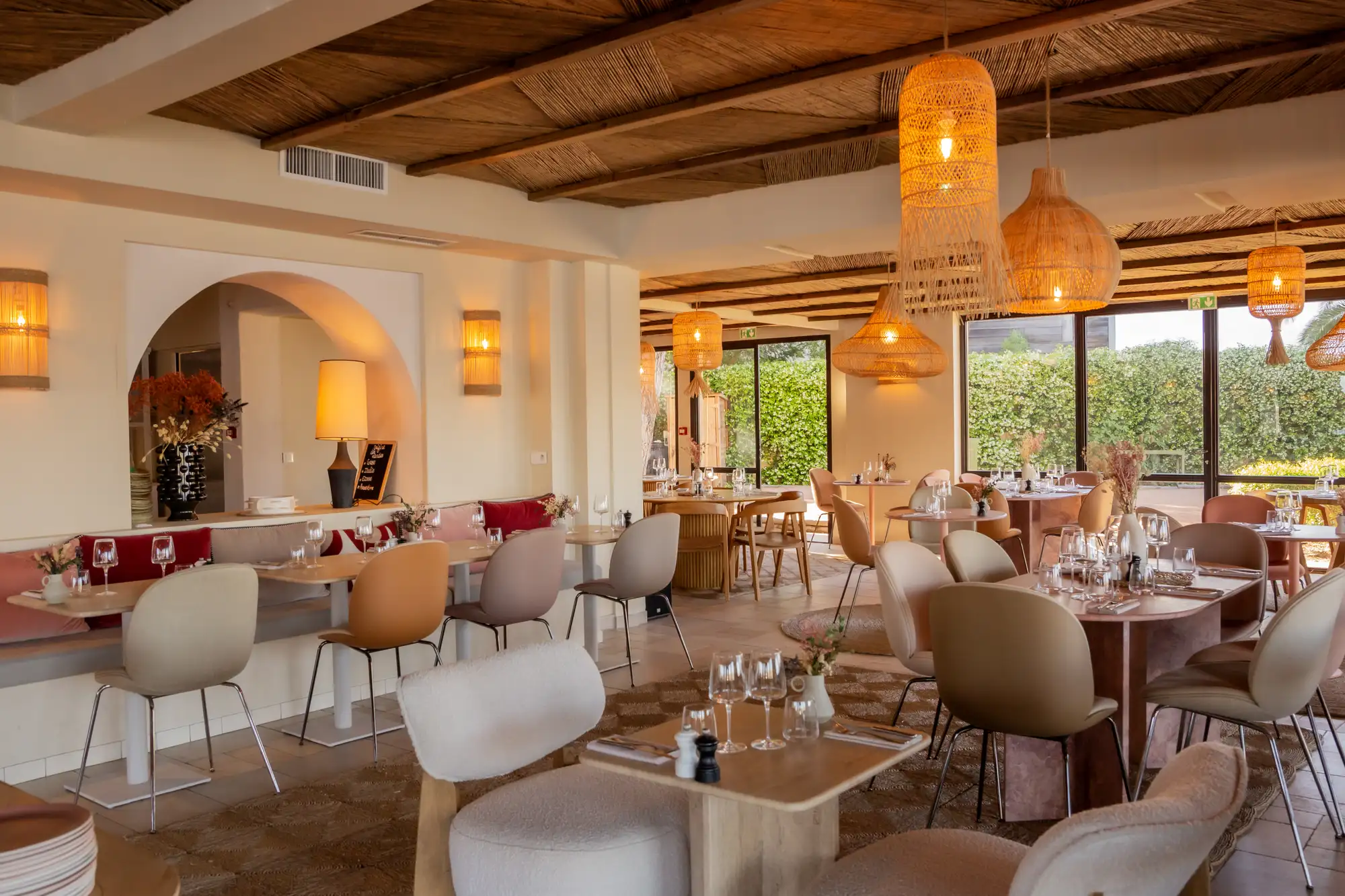 Intérieur d'un restaurant élégant avec tables en bois clair et chaises de différentes couleurs, suspensions en osier éclairées, décoration florale et grande baie vitrée donnant sur un jardin.